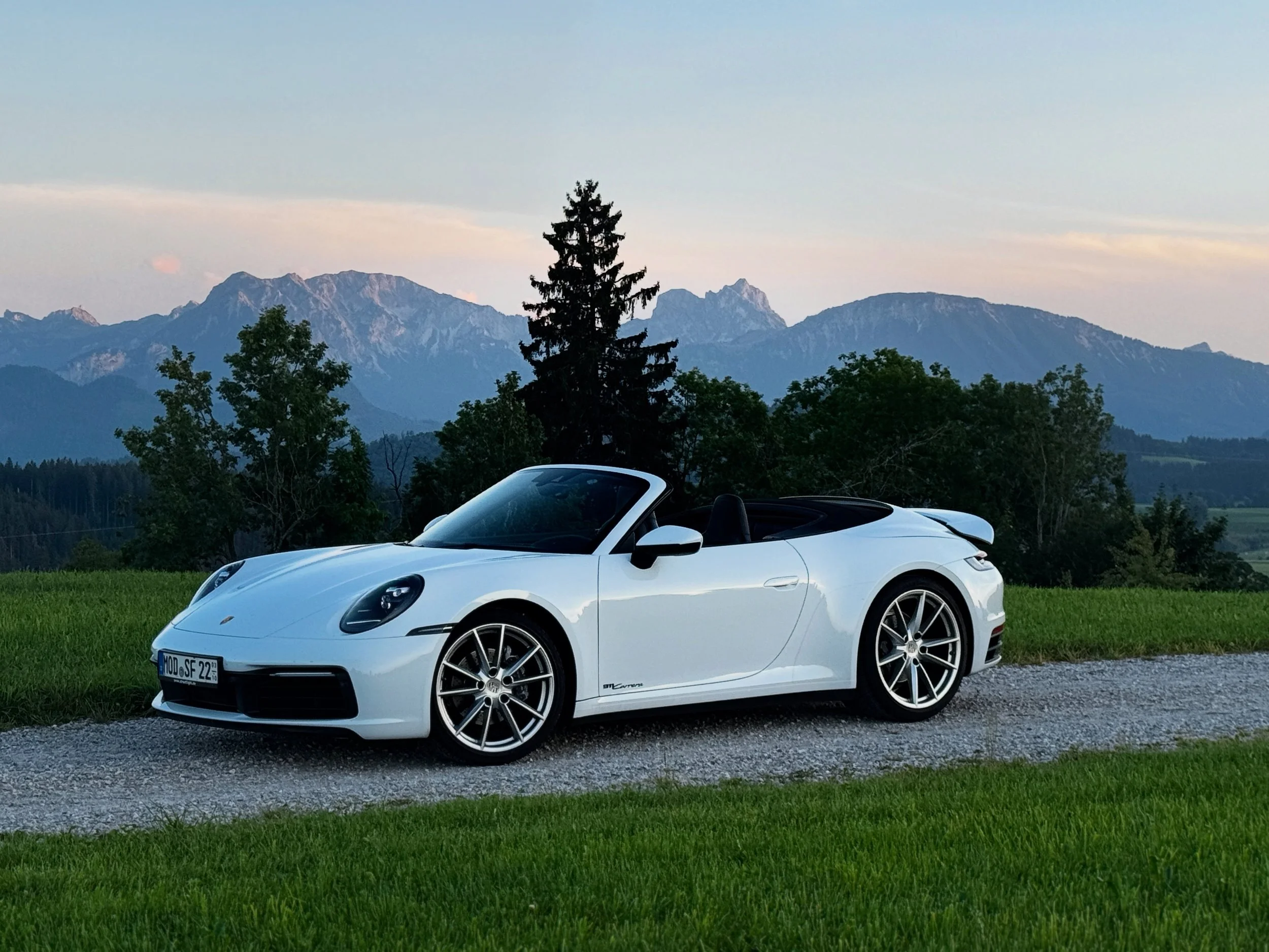 Weiße Porsche Cabriolet auf einer Wiese mit bergigem Hintergrund bei Sonnenuntergang.