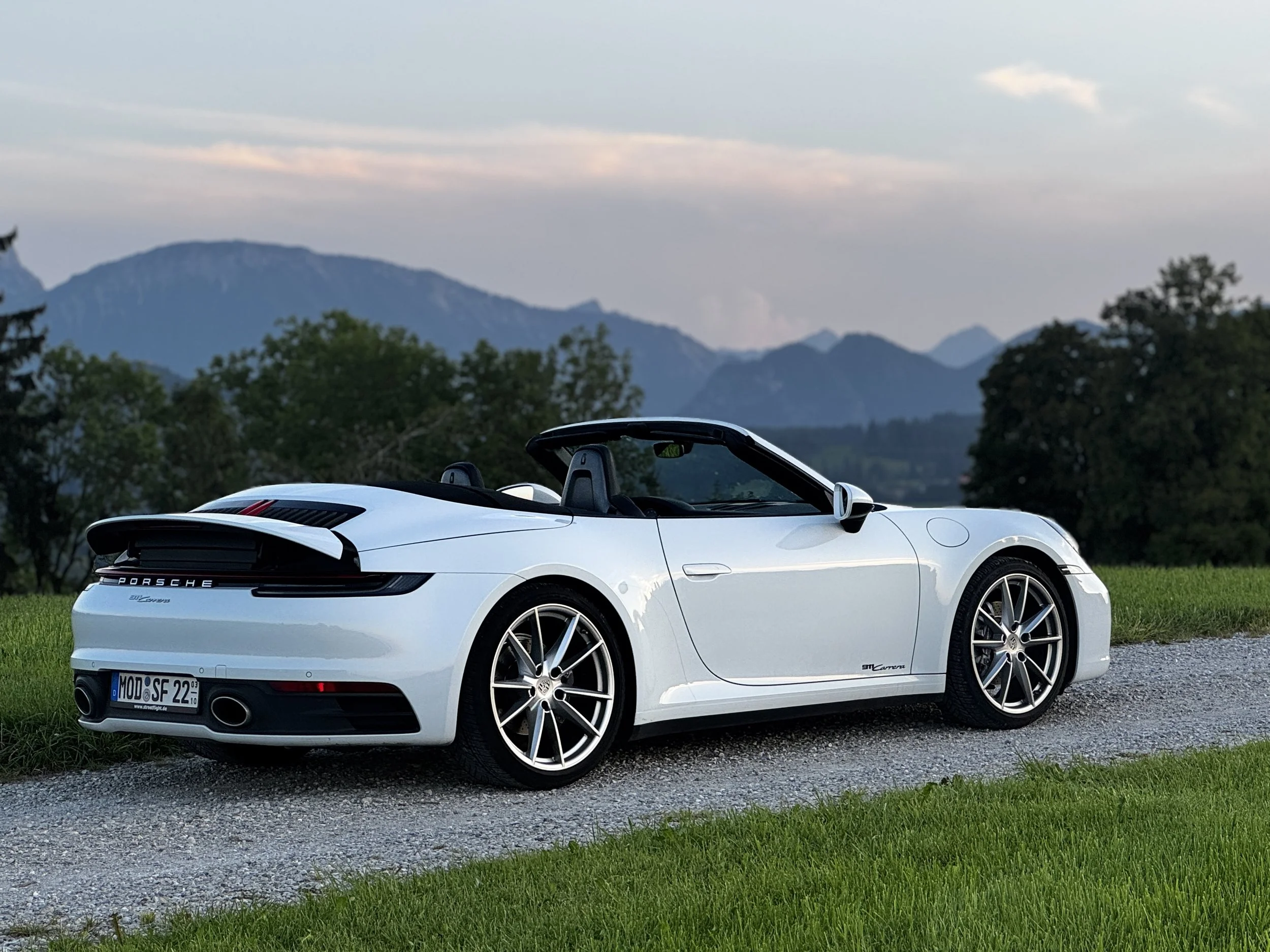 Weiße Porsche 911 Cabriolet auf einer gravel Straße, im Hintergrund Berge und Bäume bei Sonnenuntergang.