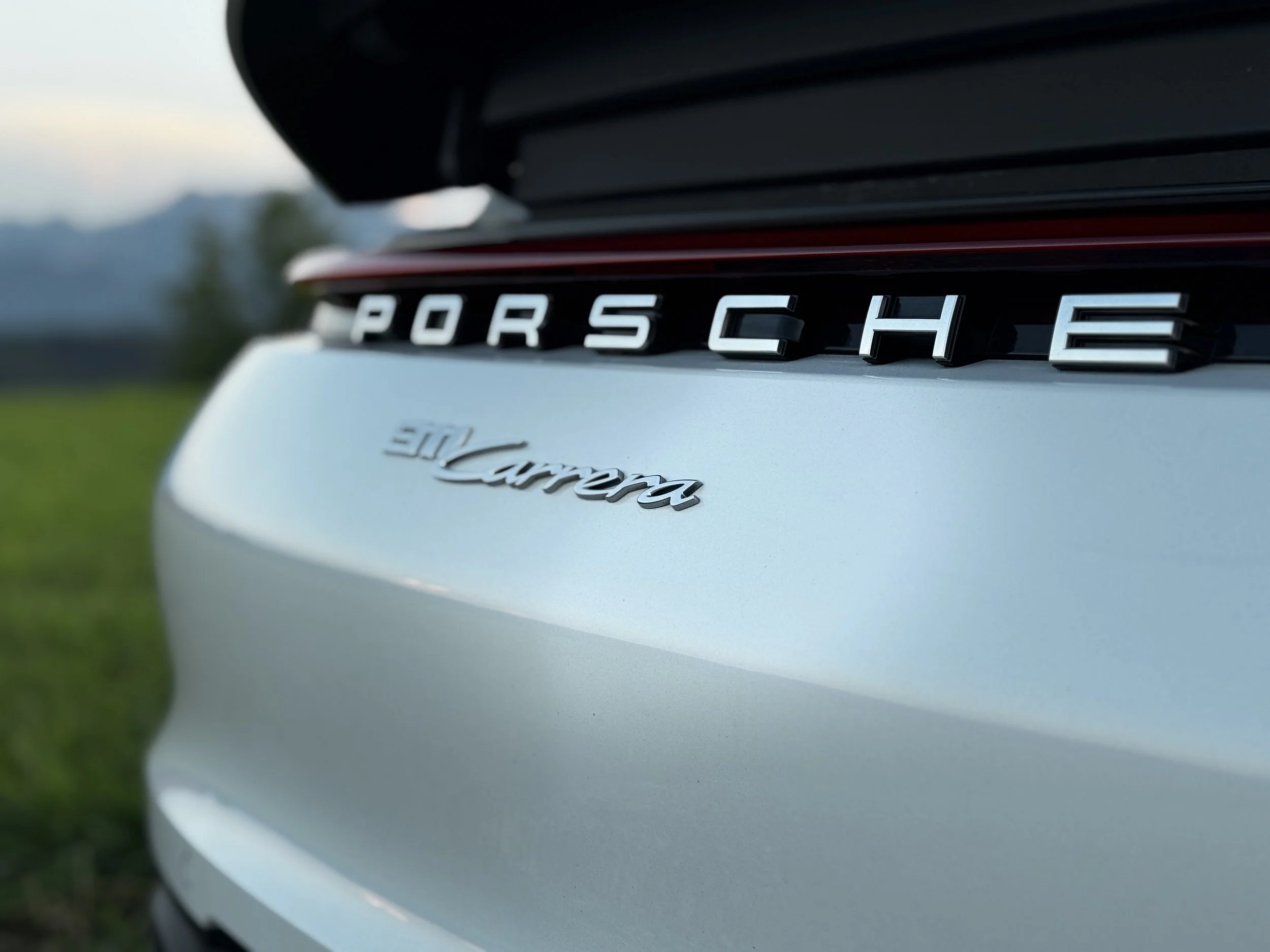 Nahaufnahme des Rückens eines weißen Porsche 911 Carrera mit Marken- und Modellbezeichnung in Silber auf der Heckklappe, im Hintergrund unscharfer Naturhintergrund.