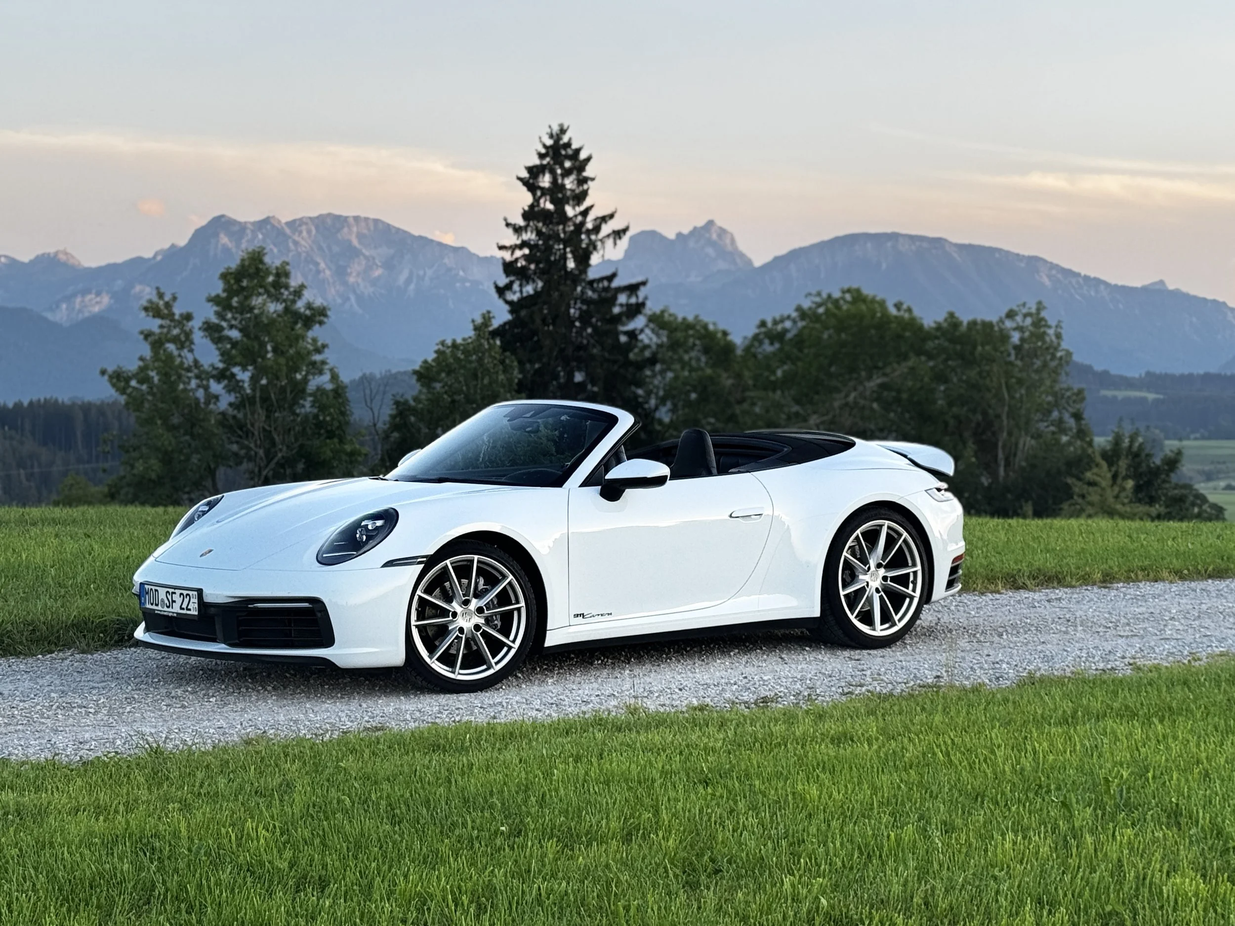 Weiße Porsche Cabrio auf einem Kiesweg, umgeben von grünem Rasen und Bäumen, im Hintergrund Berge bei Sonnenuntergang.