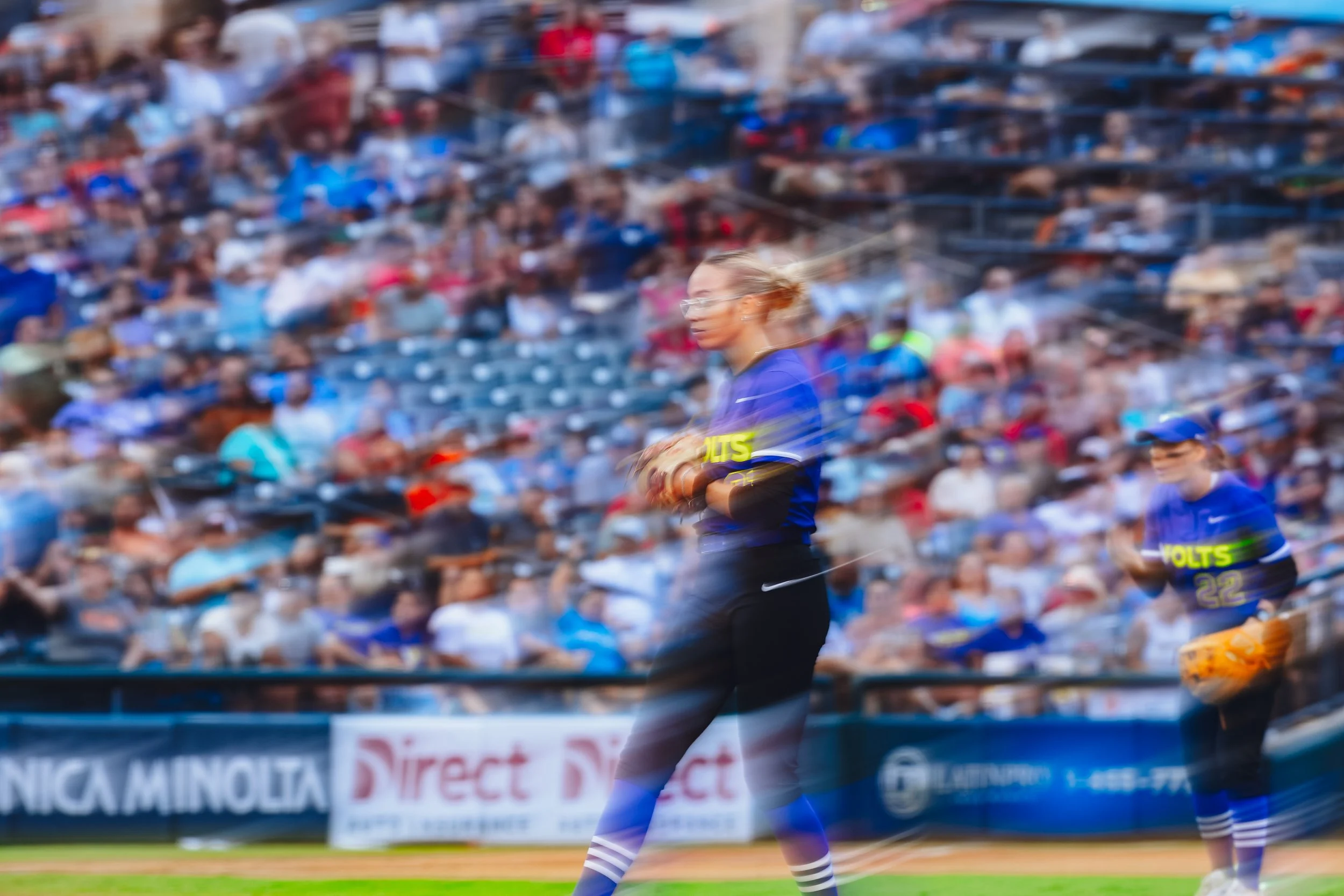Blurred image of a professional baseball game with players and a crowd in the background. A female player in a blue jersey and black pants is in the foreground, holding a baseball glove.
