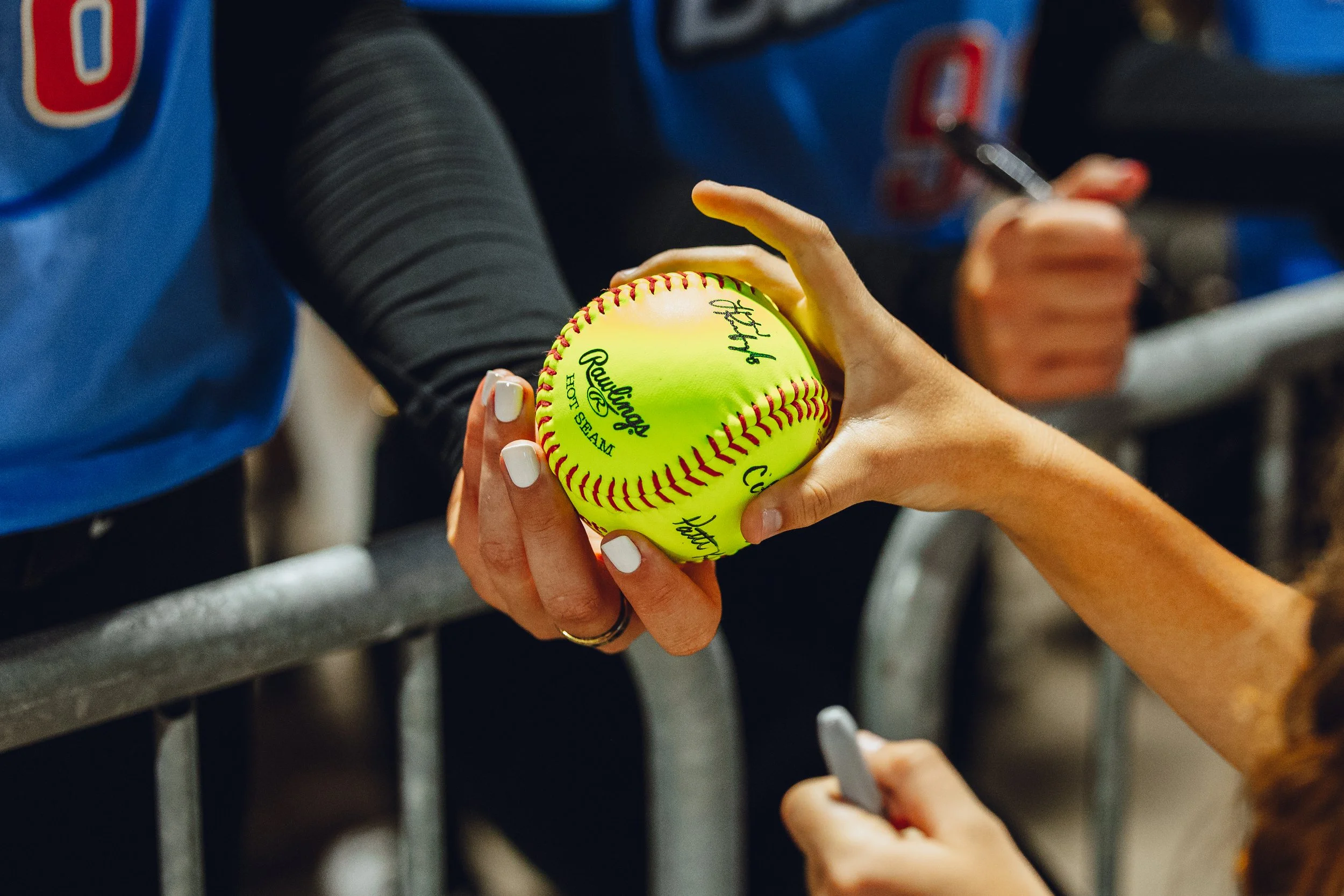 A person signing a bright yellow softball with red stitching, titled 'Rawlings' and 'Official' during a signing event.