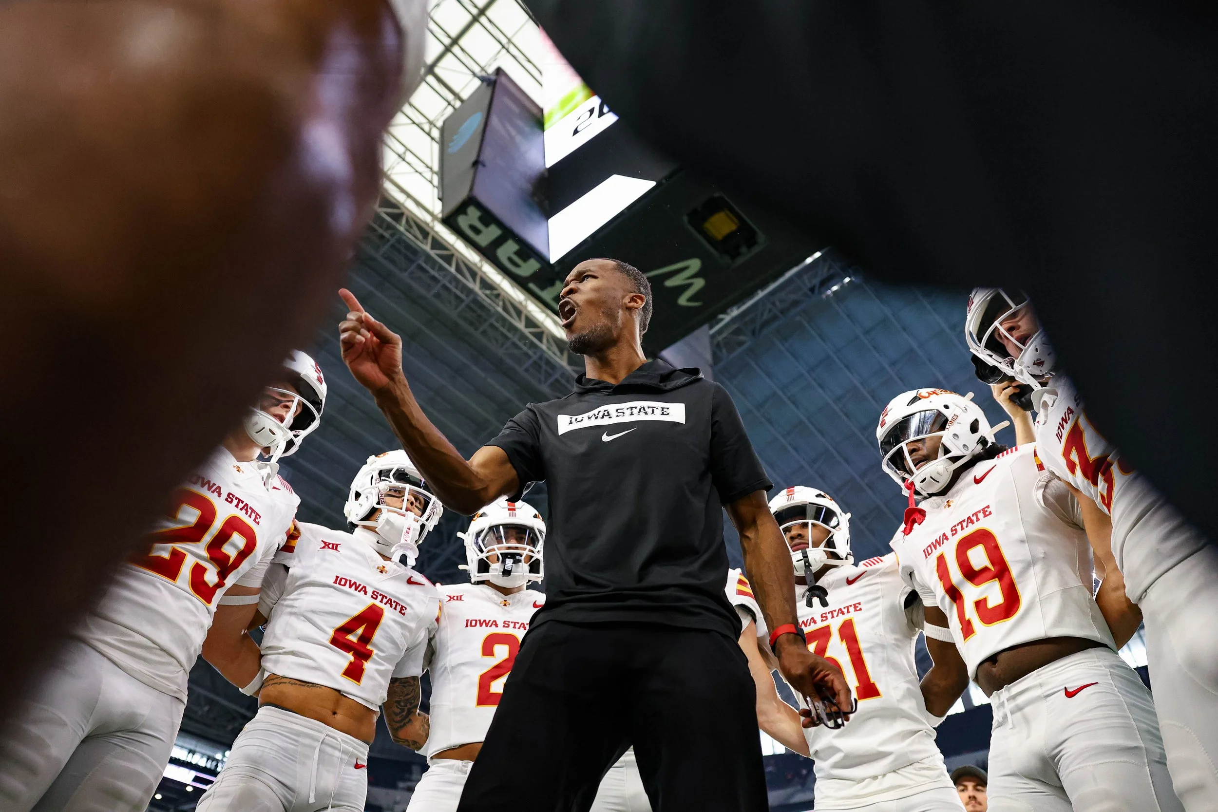 A coach, wearing a black shirt that says 'Iowa State', is passionately speaking to a group of Iowa State football players who are dressed in white uniforms with red and yellow accents inside a stadium.