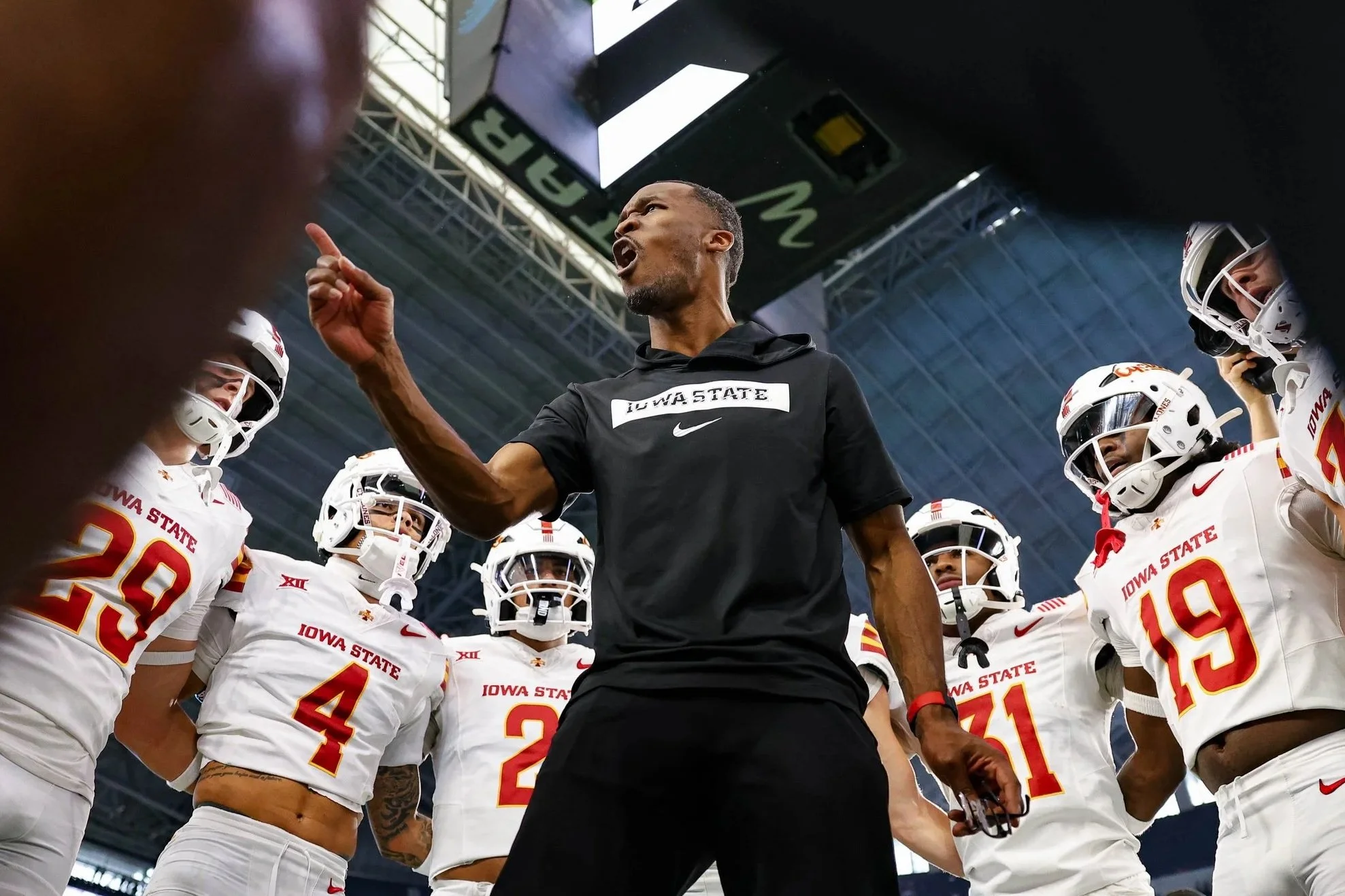 A coach, wearing a black shirt that says 'Iowa State', is passionately speaking to a group of Iowa State football players who are dressed in white uniforms with red and yellow accents inside a stadium.