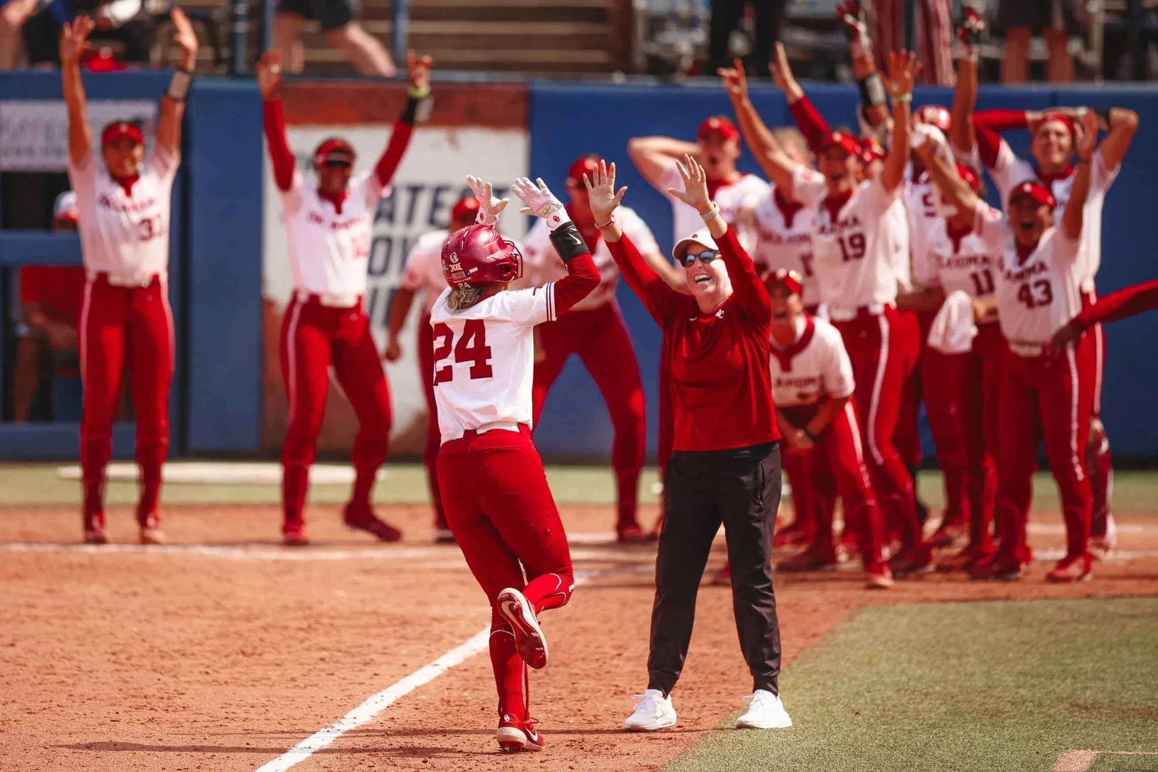 A female softball player high-fives a woman, possibly her coach, on the field near home plate during a game. In the background, a group of players and staff cheer and celebrate.