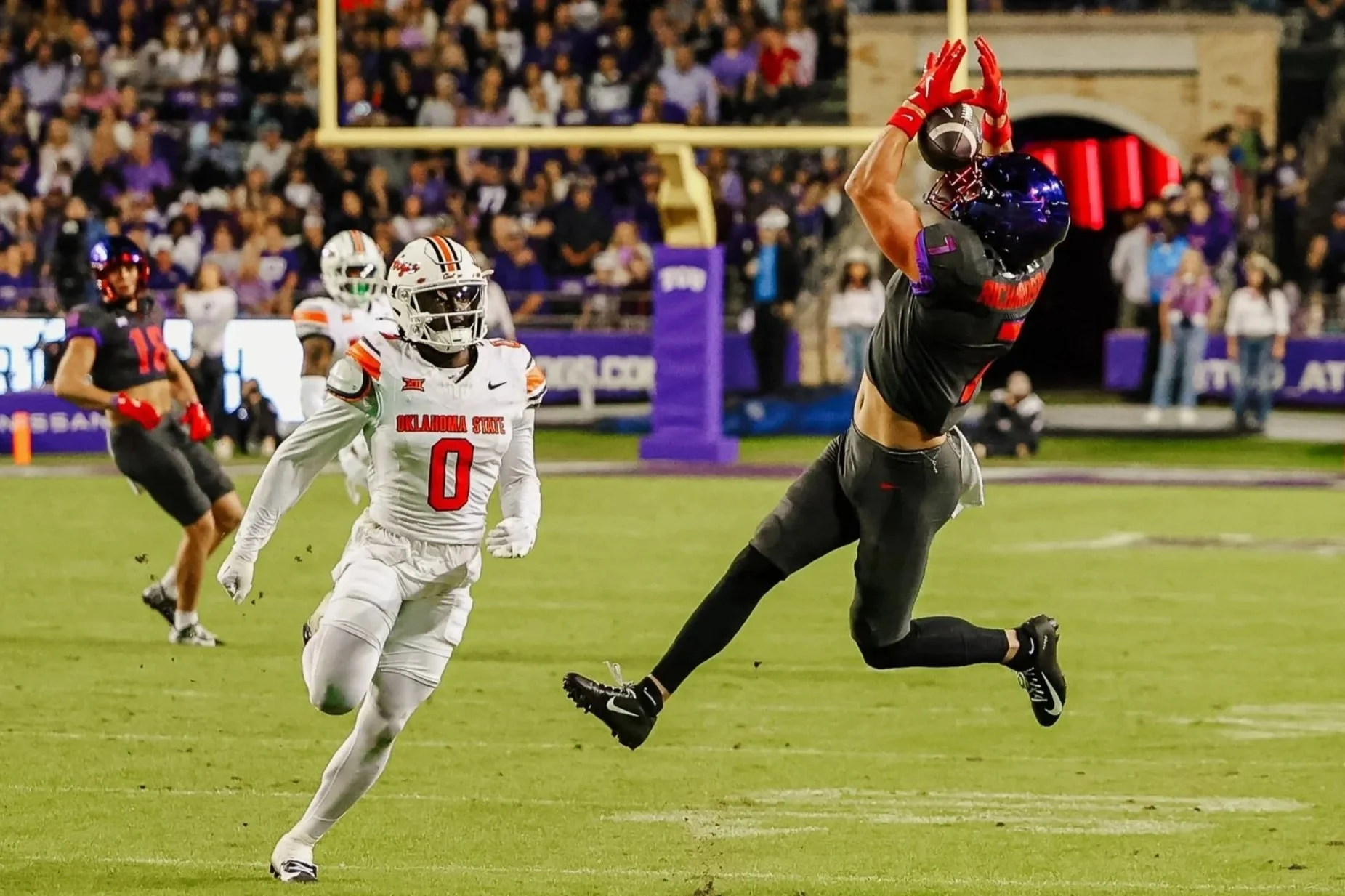 A football player in black uniform jumping to catch a football in the end zone while being guarded by a player in white uniform with number 0 on the jersey. Other players and spectators are visible in the background.