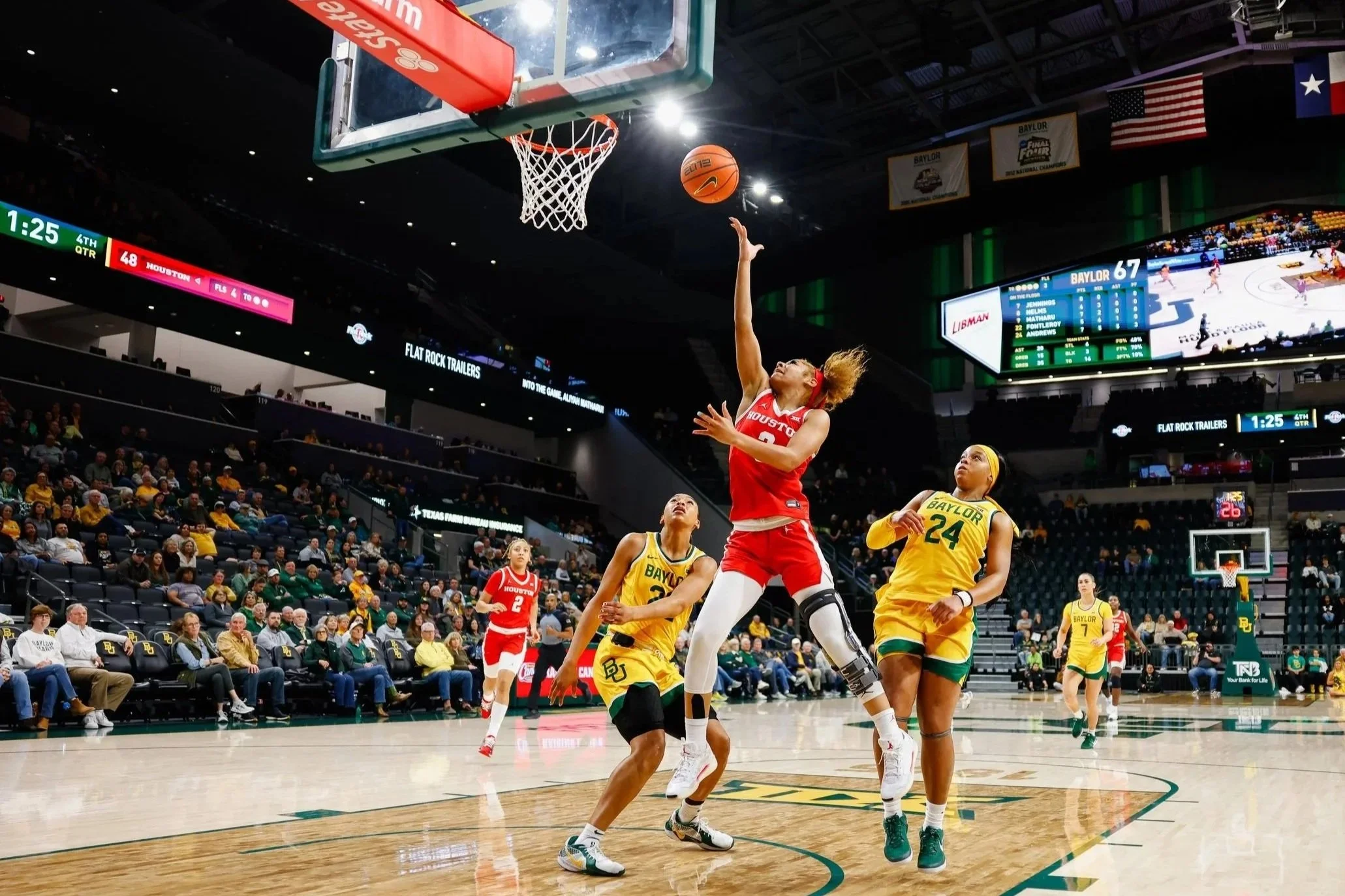 A women's basketball game between Houston and Baylor, with players on the court, one player in red jumping to score or block, and others in yellow defending; audience seated in the stands; scoreboard displaying Houston leading Baylor.