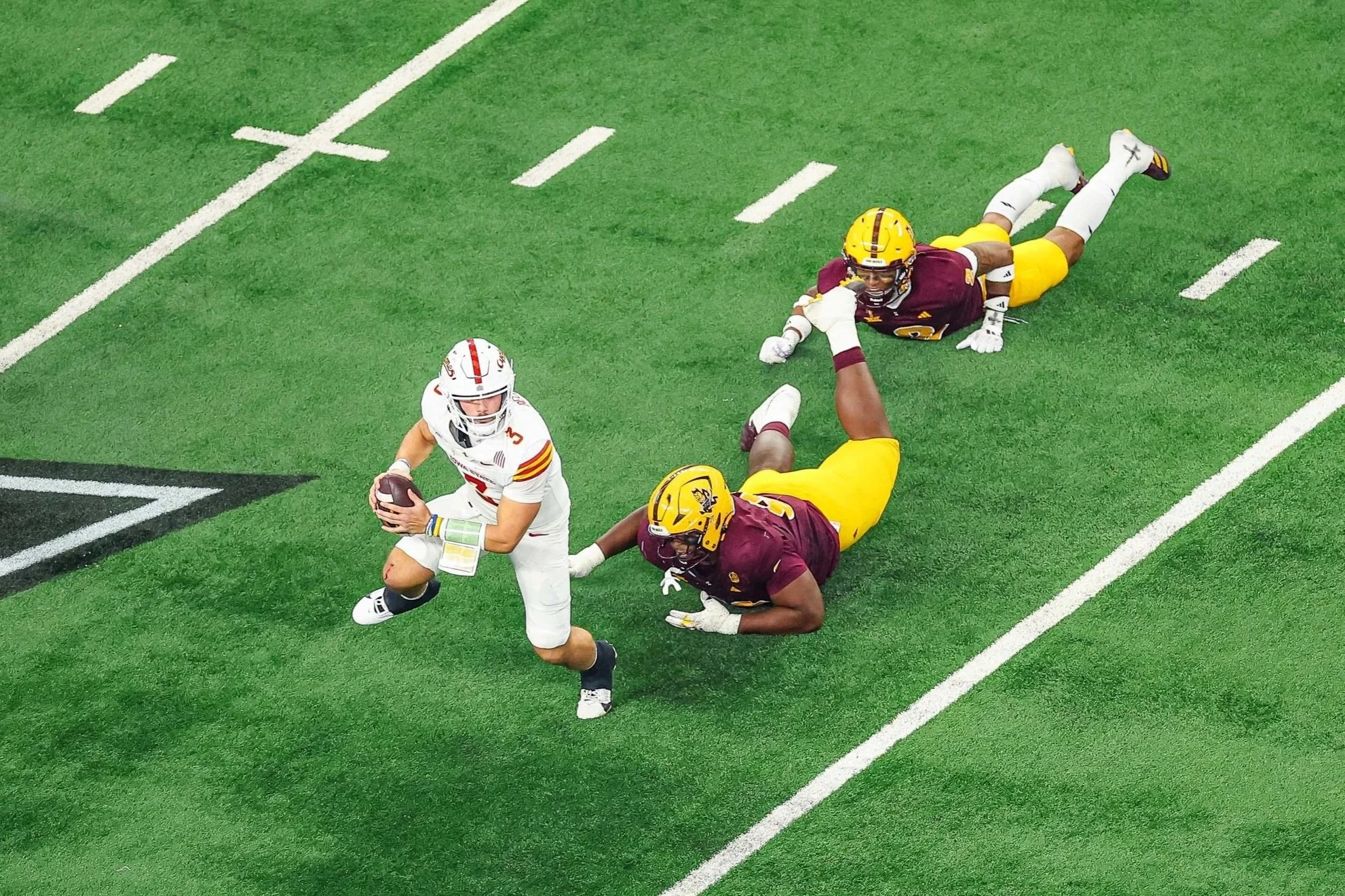 A football player in white uniform holding a football runs while two players in maroon and yellow uniforms try to tackle him on a green field.
