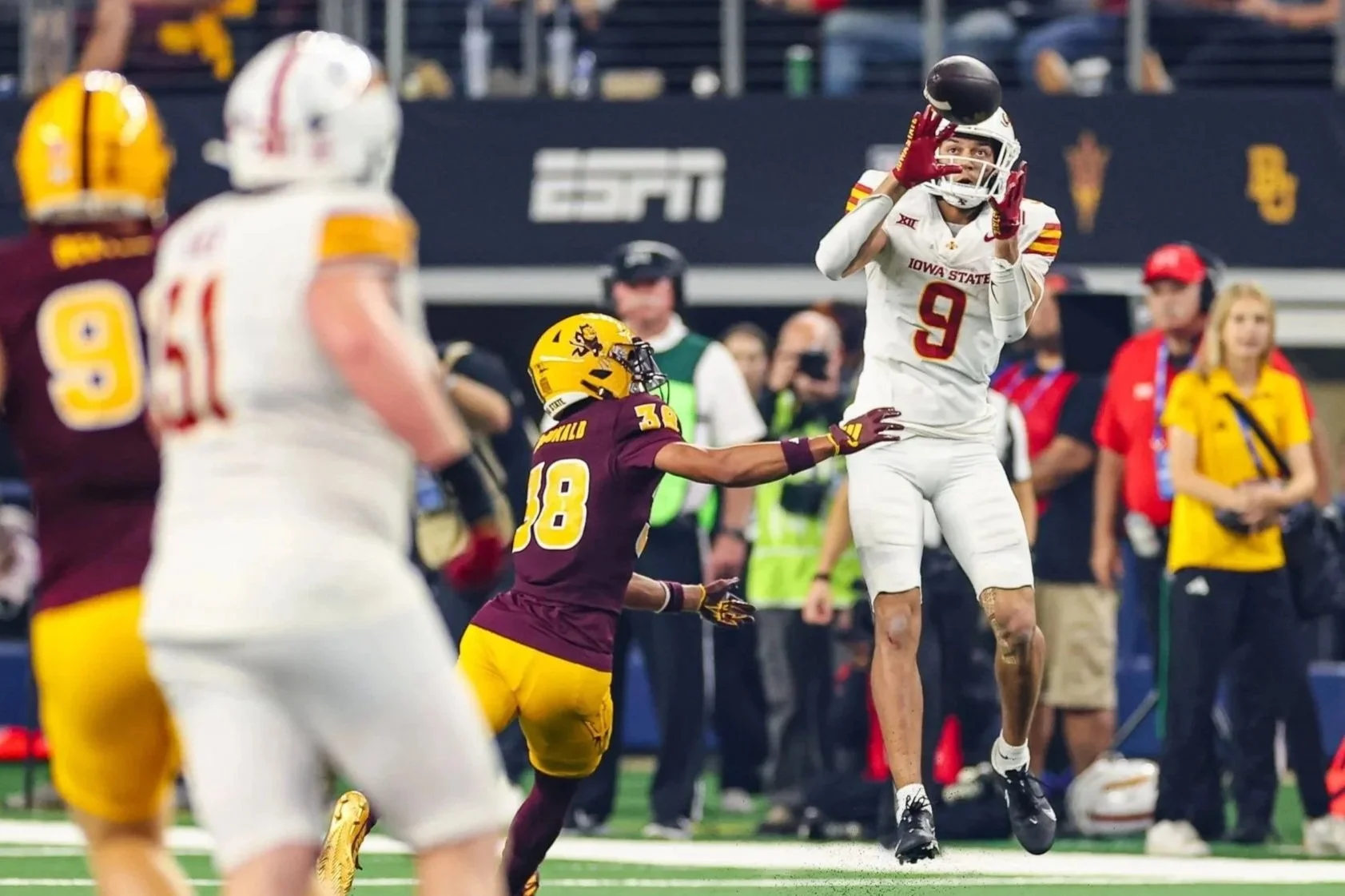 Iowa State football player in white uniform catching football while opponent in maroon and yellow reaches to defend, on field at stadium with spectators in background.