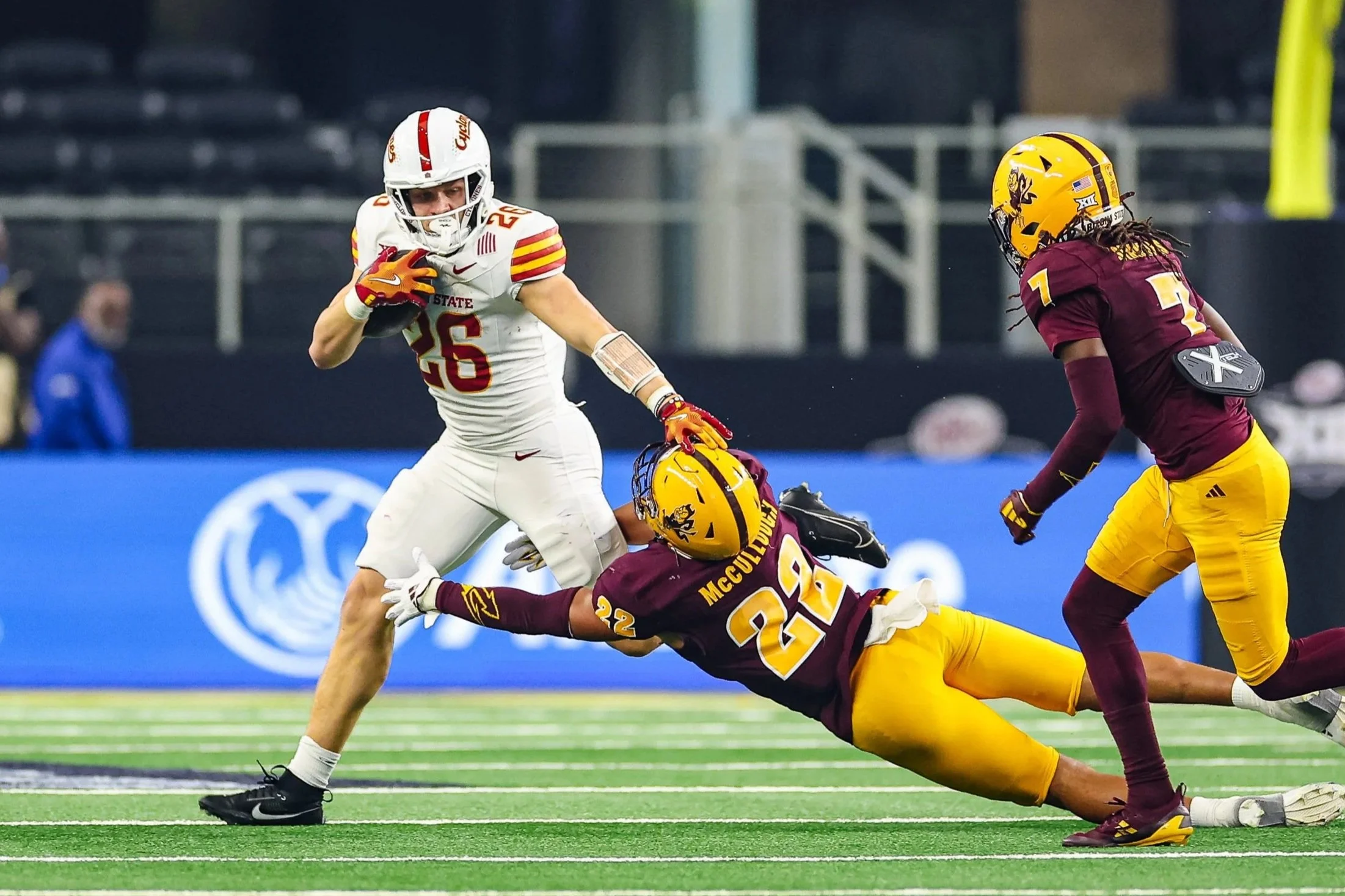 A football game with a player in a white uniform number 26 running with the ball while two players in maroon and yellow uniforms attempt to tackle him on a green field inside a stadium.