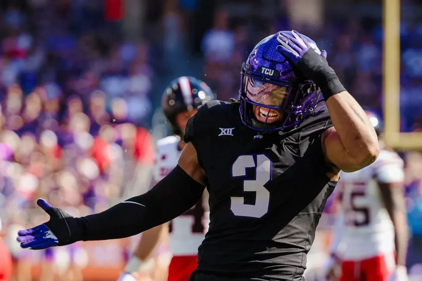 A football player in a black jersey with the number 3, wearing purple pants and a purple helmet, celebrating on the field with a smile and a hand on his head during a game. Other players and a crowd are visible in the background.