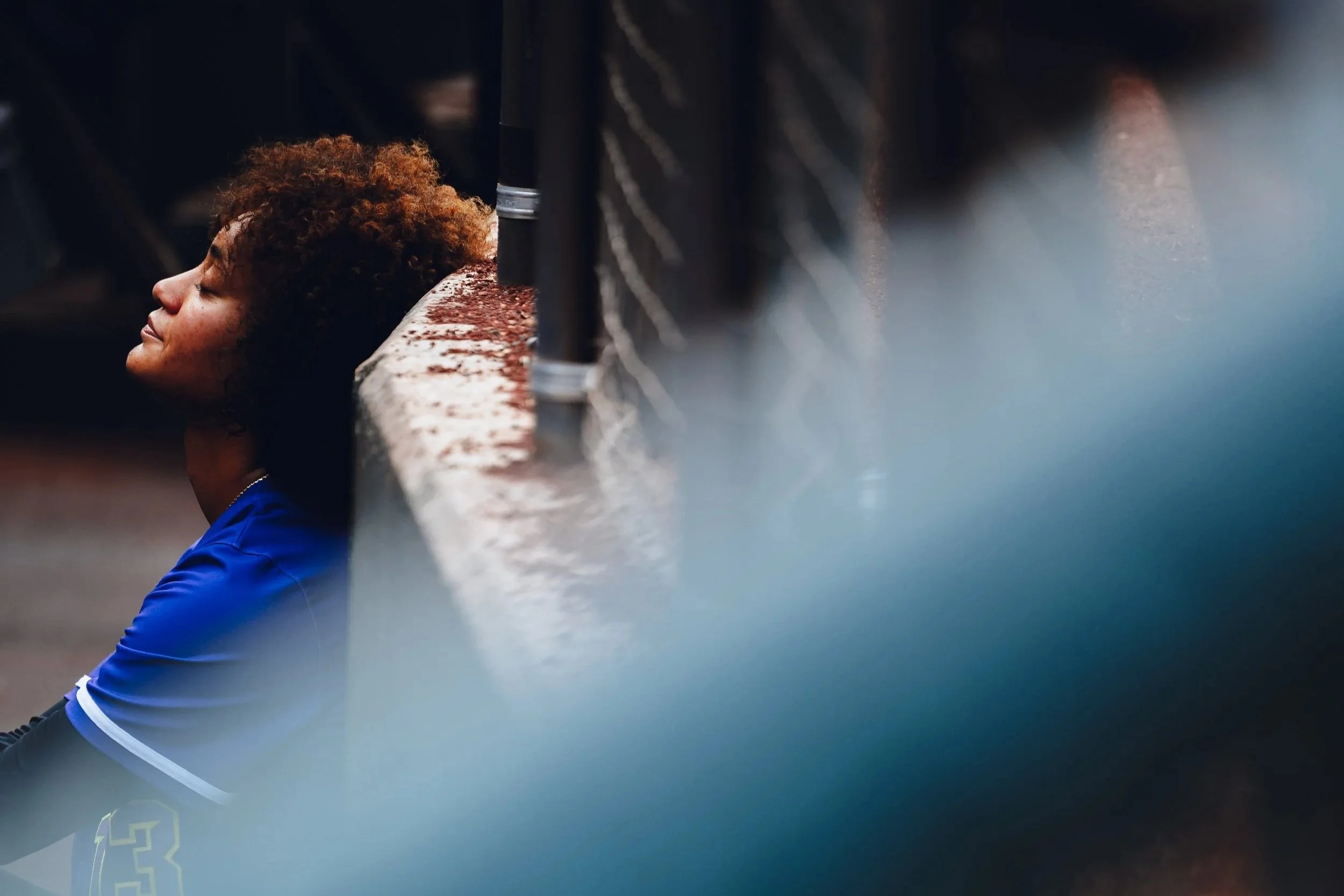 A woman with curly hair is sitting on the ground, leaning against a concrete wall with her eyes closed.
