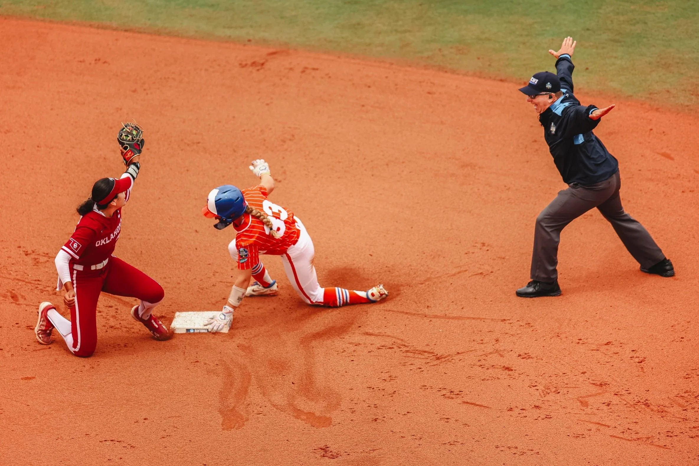 Young baseball player in a red and orange uniform slides into a base as an umpire calls the play, while a teammate in a red uniform knees nearby on the reddish dirt field.