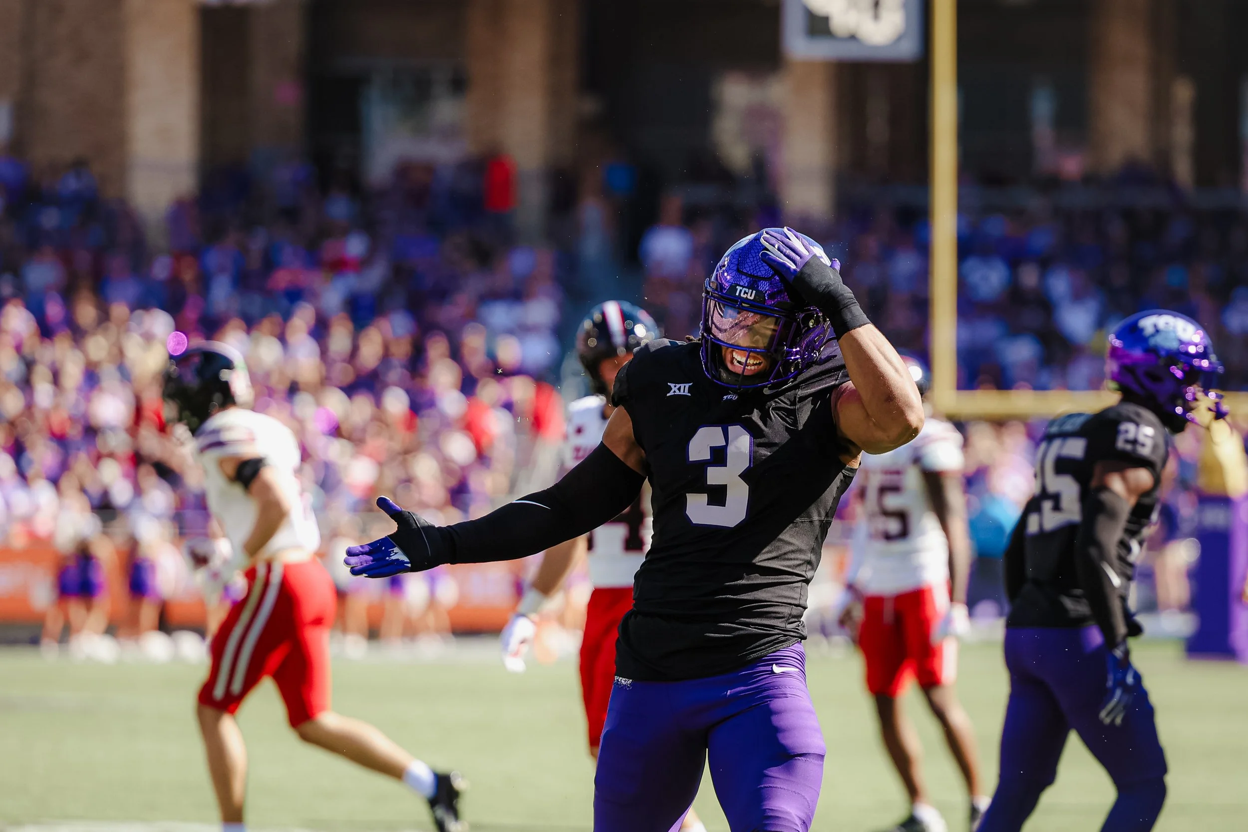 A football player in a black jersey with the number 3, wearing purple pants and a purple helmet, celebrating on the field with a smile and a hand on his head during a game. Other players and a crowd are visible in the background.
