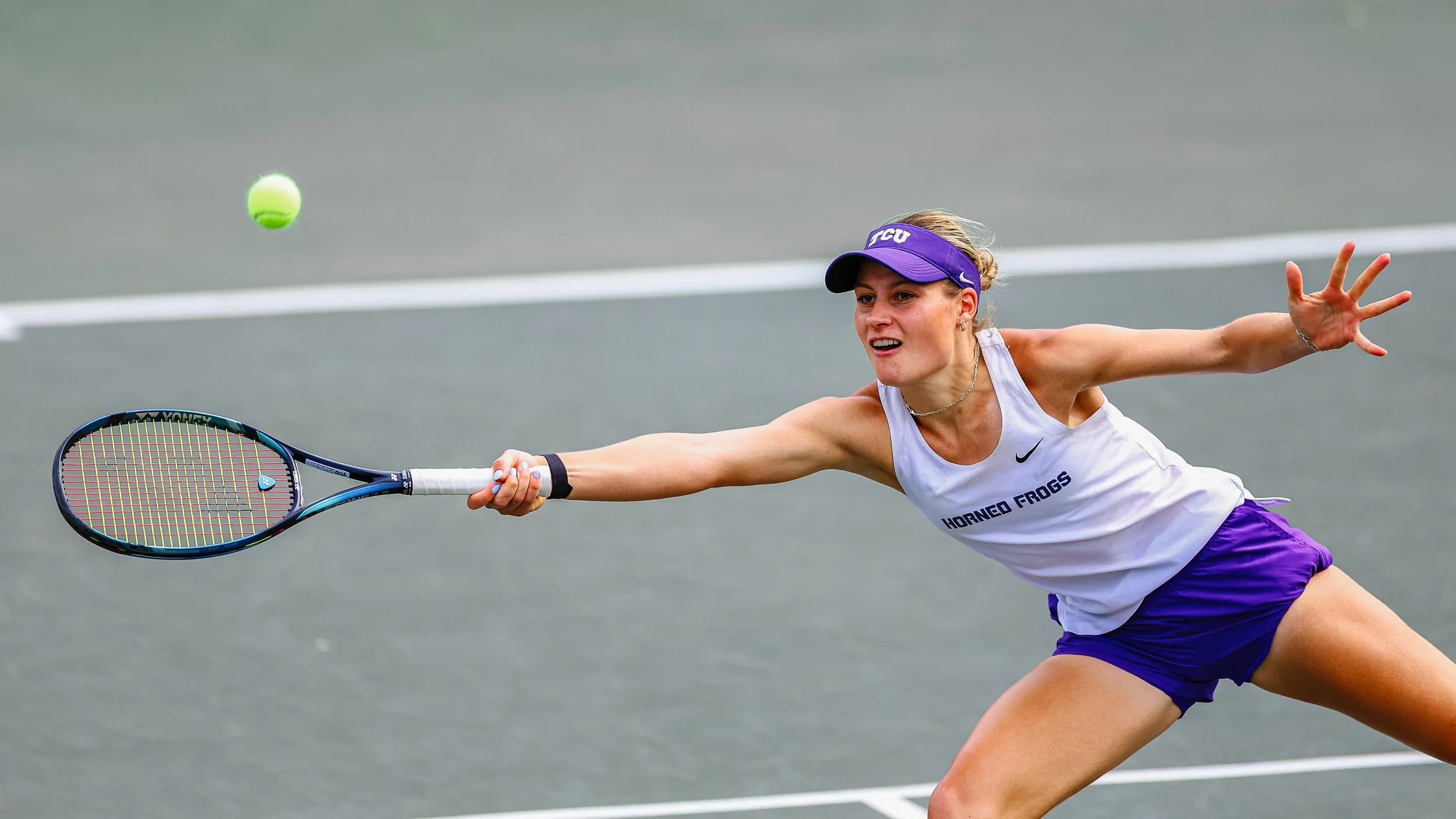 A woman playing tennis on a court, wearing a white tank top with 'HORNEO FROGS' and purple shorts, reaching to hit a yellow tennis ball with her racket, wearing a purple visor.