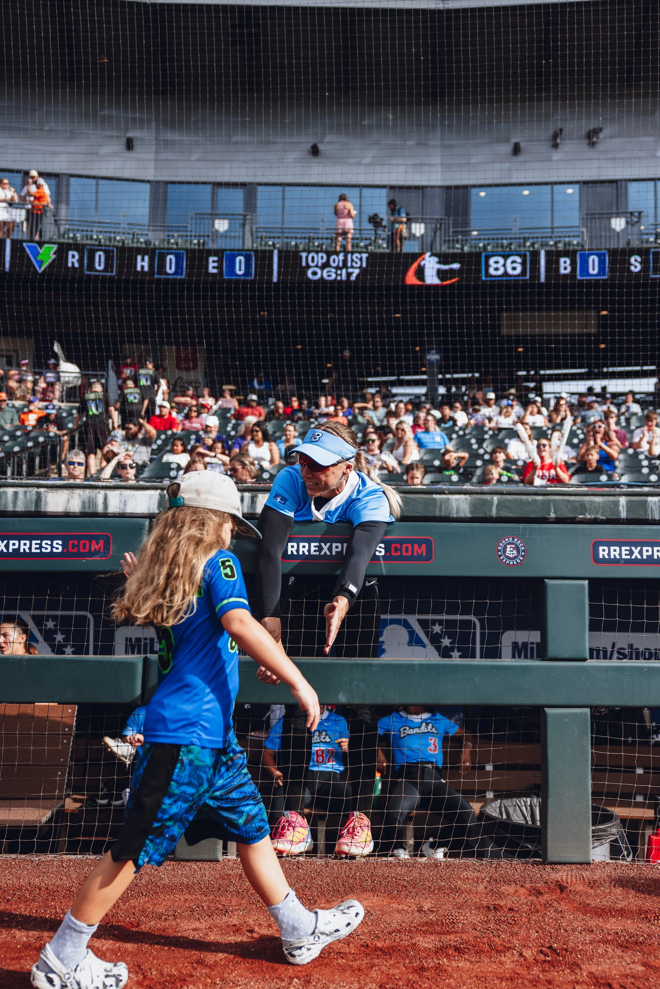 A young girl and a woman at a baseball game, with the girl walking past the woman in the dugout. The woman is leaning over the dugout railing, smiling, and reaching out towards the girl. The stadium is filled with spectators, and an electronic scoreb