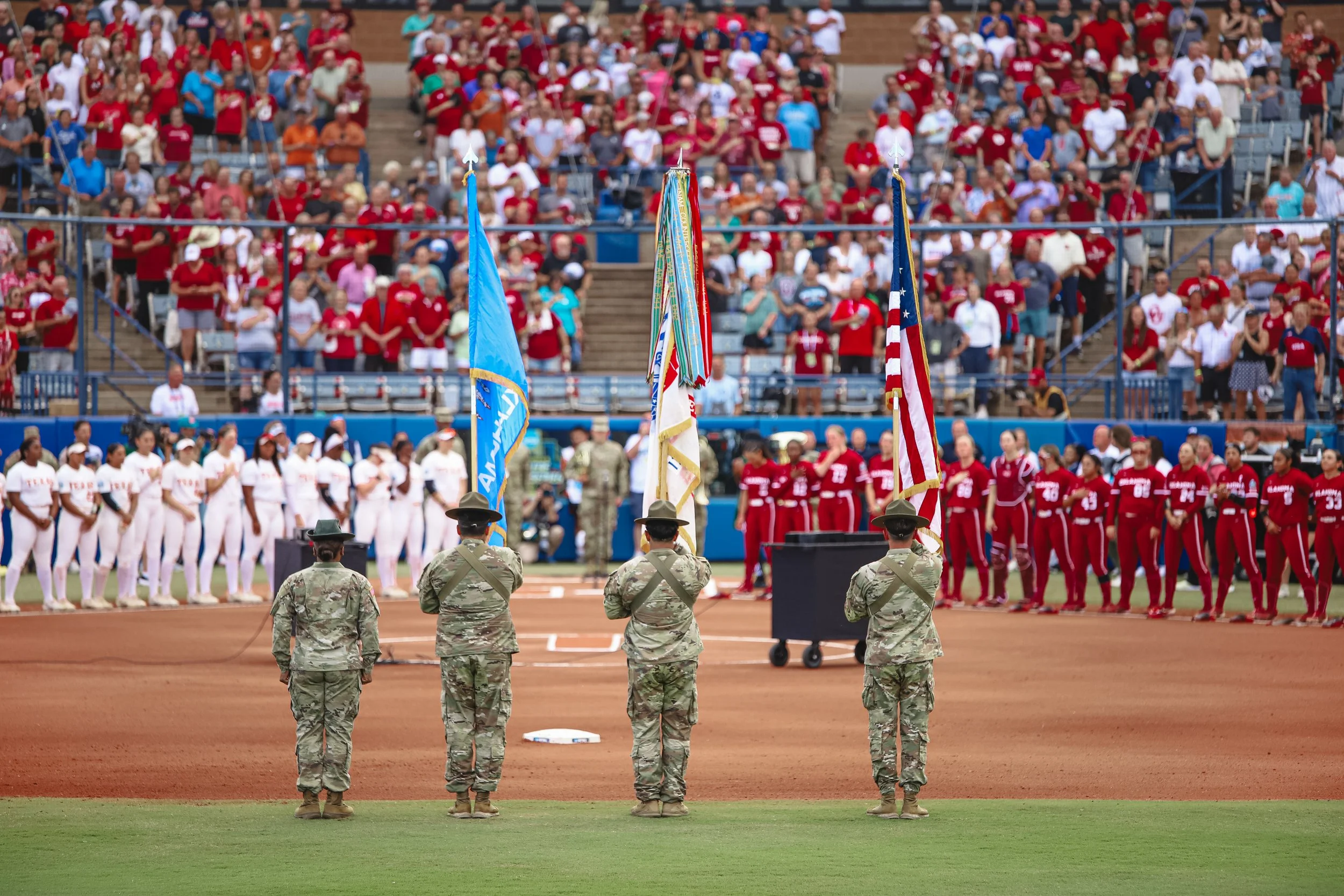 Soldiers holding flags at a patriotic event on a baseball field with a large crowd in the stands, including teams dressed in white and red uniforms.