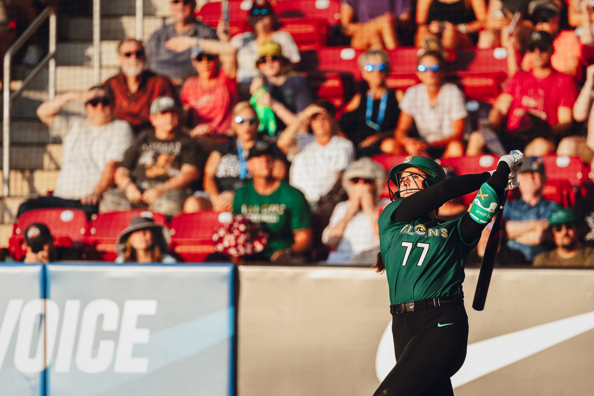 A female softball player wearing a green uniform with the number 77 is swinging a bat on a baseball field. Spectators sit in the background in red stadium seats, some wearing sunglasses and hats, watching the game.