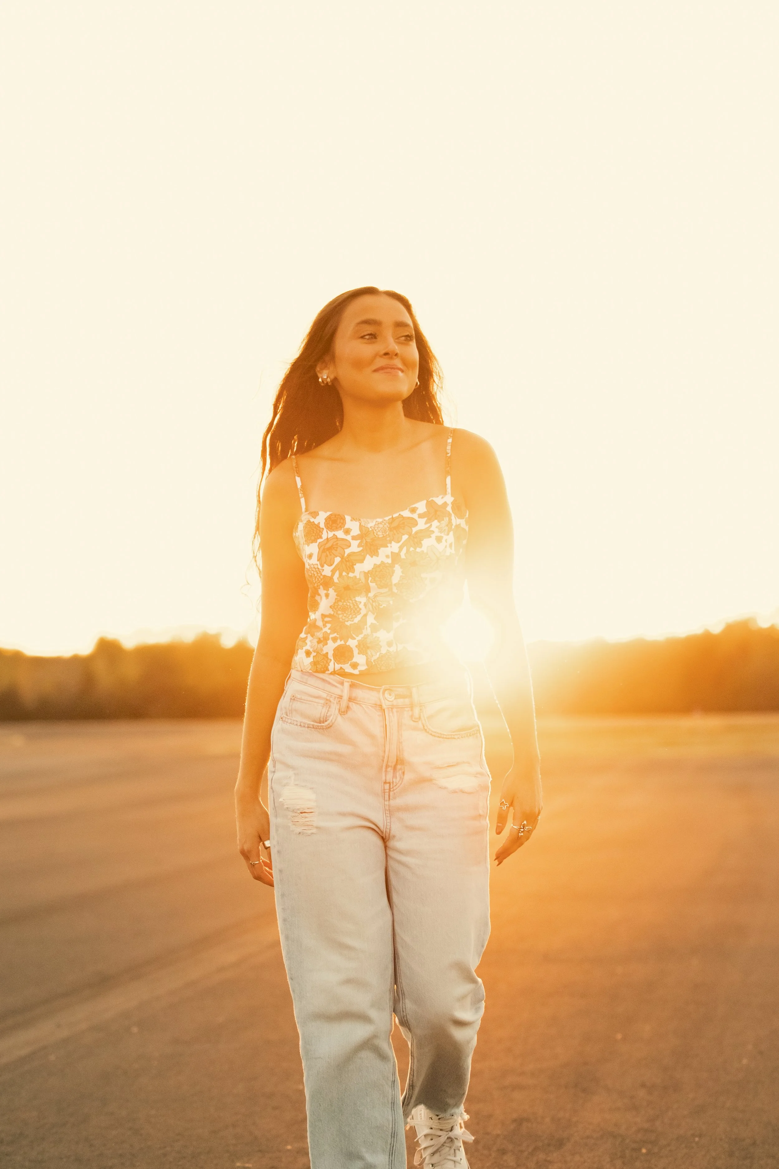 A woman walking outdoors at sunset, wearing a floral top and jeans, with the sun behind her.