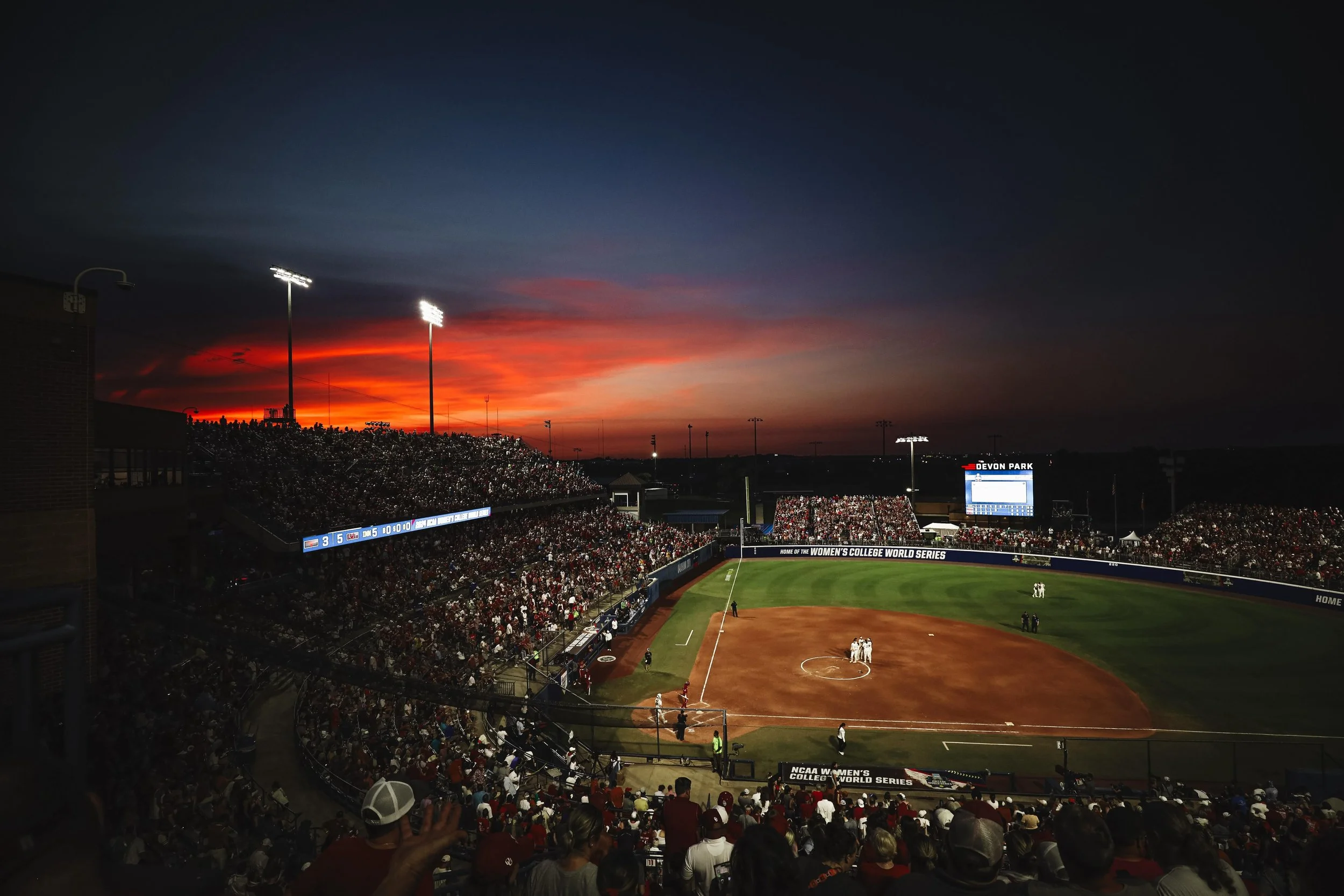 A baseball stadium filled with spectators during sunset, with a vibrant red and orange sky in the background.