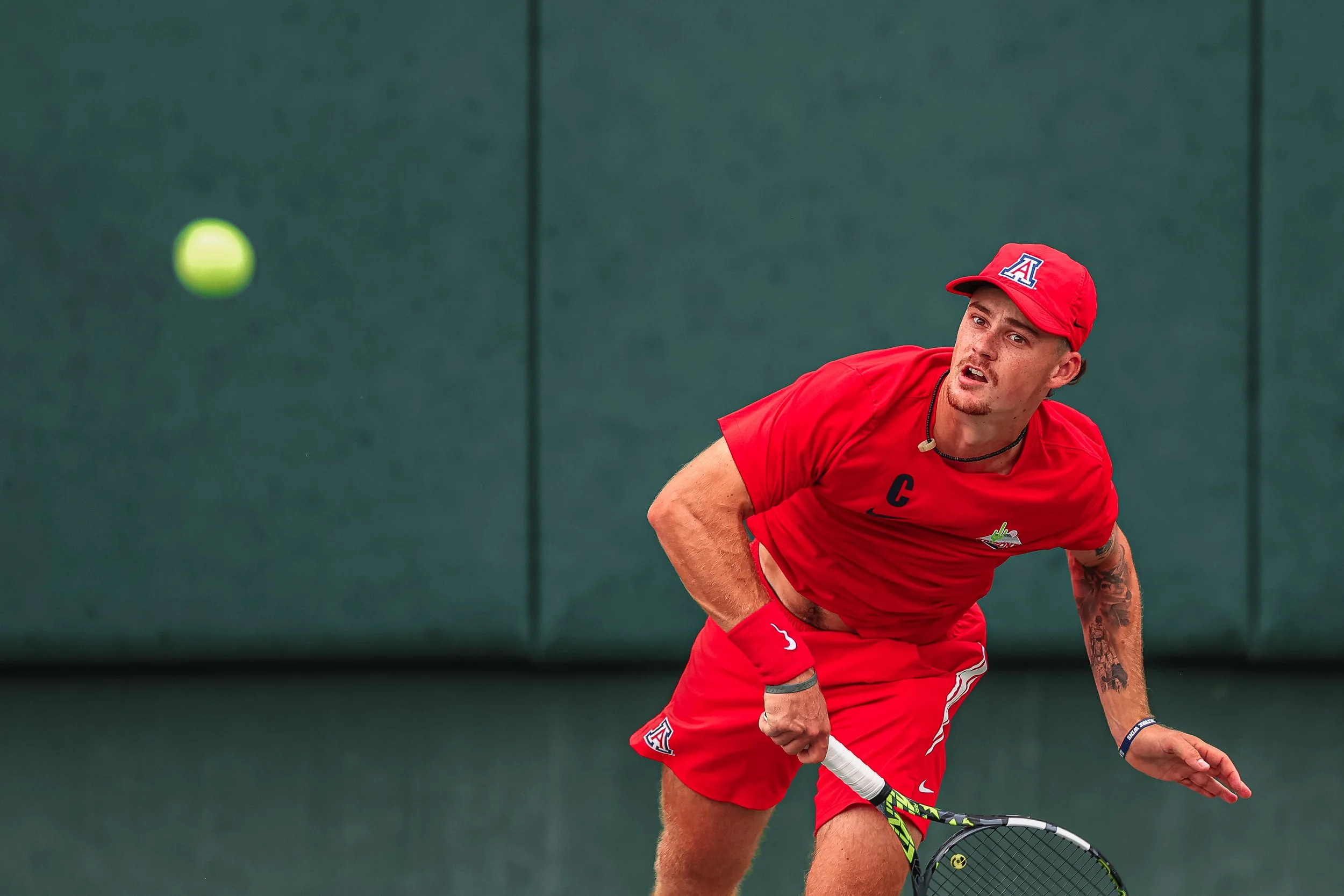 A man in a red tennis outfit with a cap, holding a tennis racket, is chasing a tennis ball on a green court.