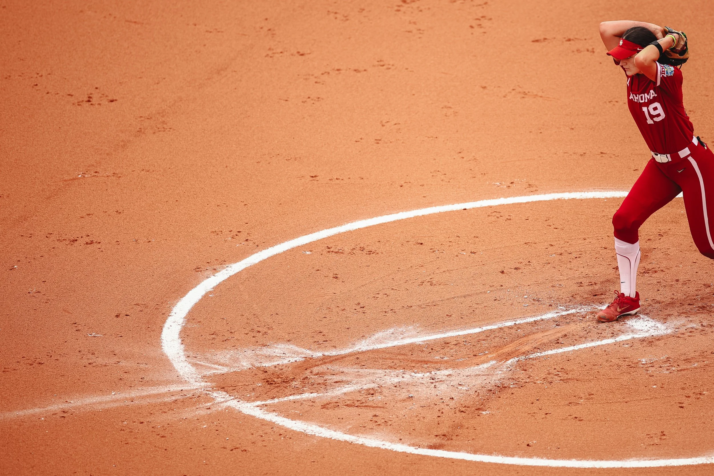 A female softball player in a red uniform with the number 19 on the jersey, standing on a dirt field near home plate, preparing to throw or catch a ball.
