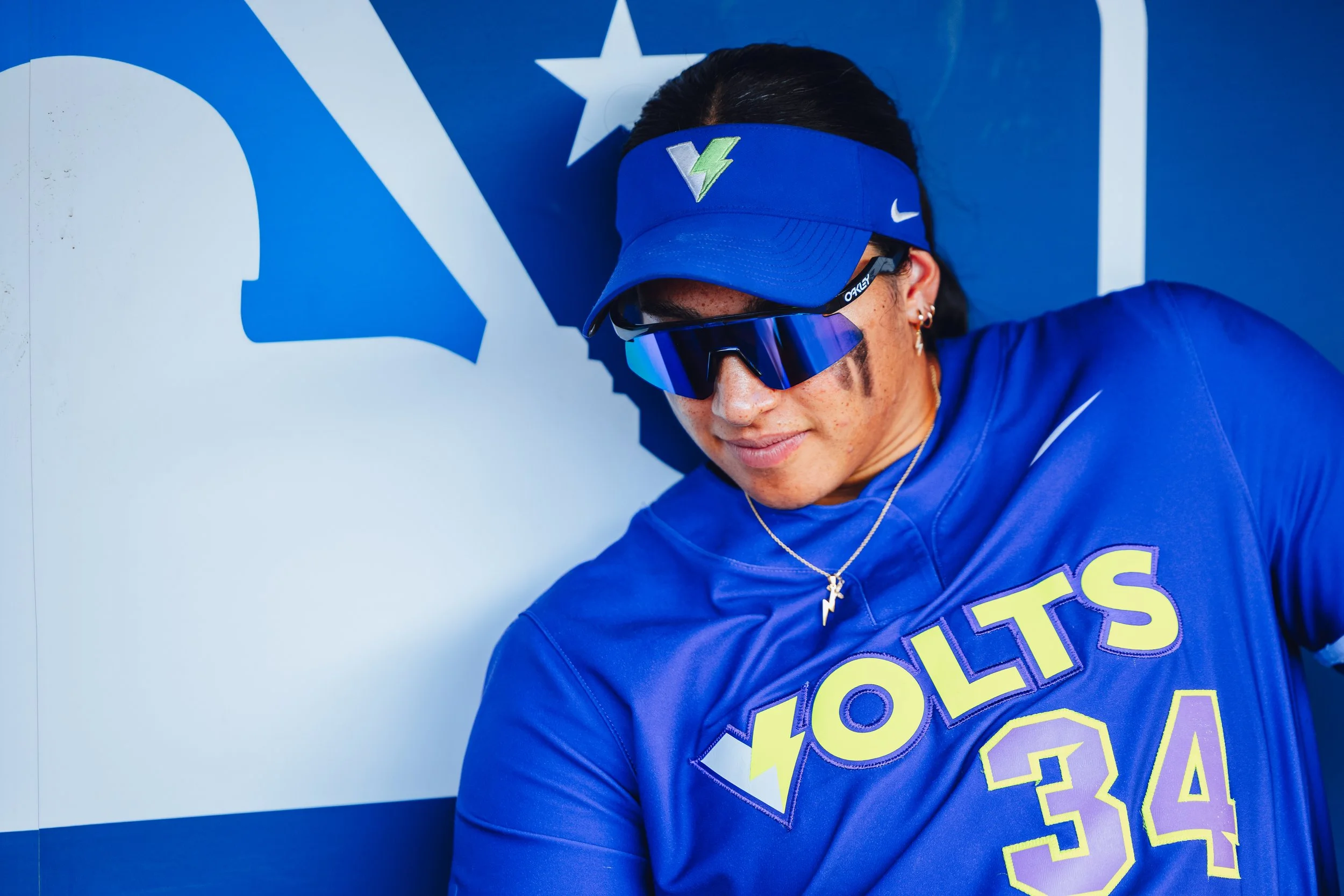 A female athlete dressed in a blue sports jersey and visor, wearing sunglasses and jewelry, leaning against a wall with the word 'VOLT' and the number 34.