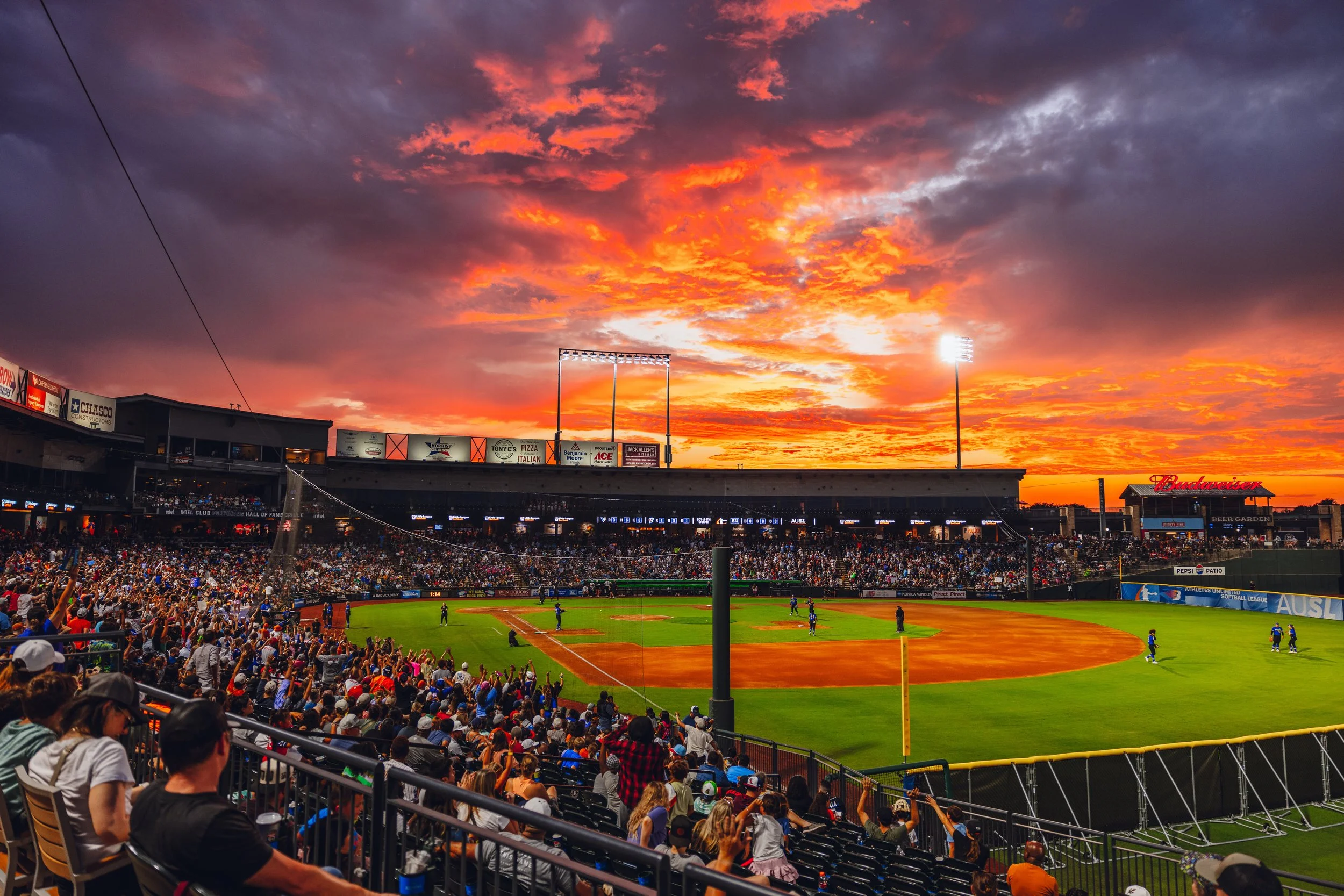 Crowd of baseball fans watching a game at a stadium during sunset with a vibrant orange and purple sky.