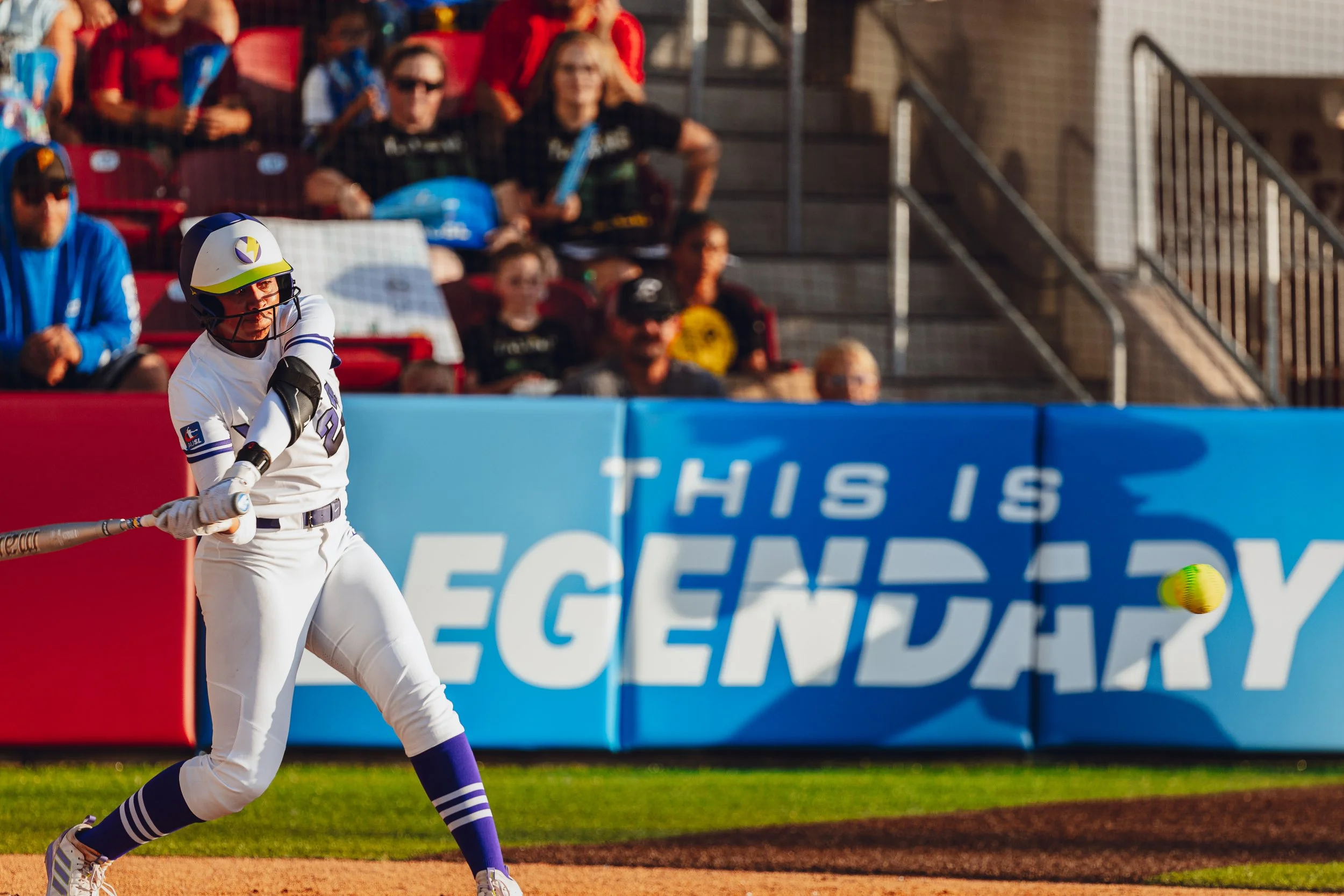 A baseball player swings a bat during a game, with spectators watching from the stands in the background.