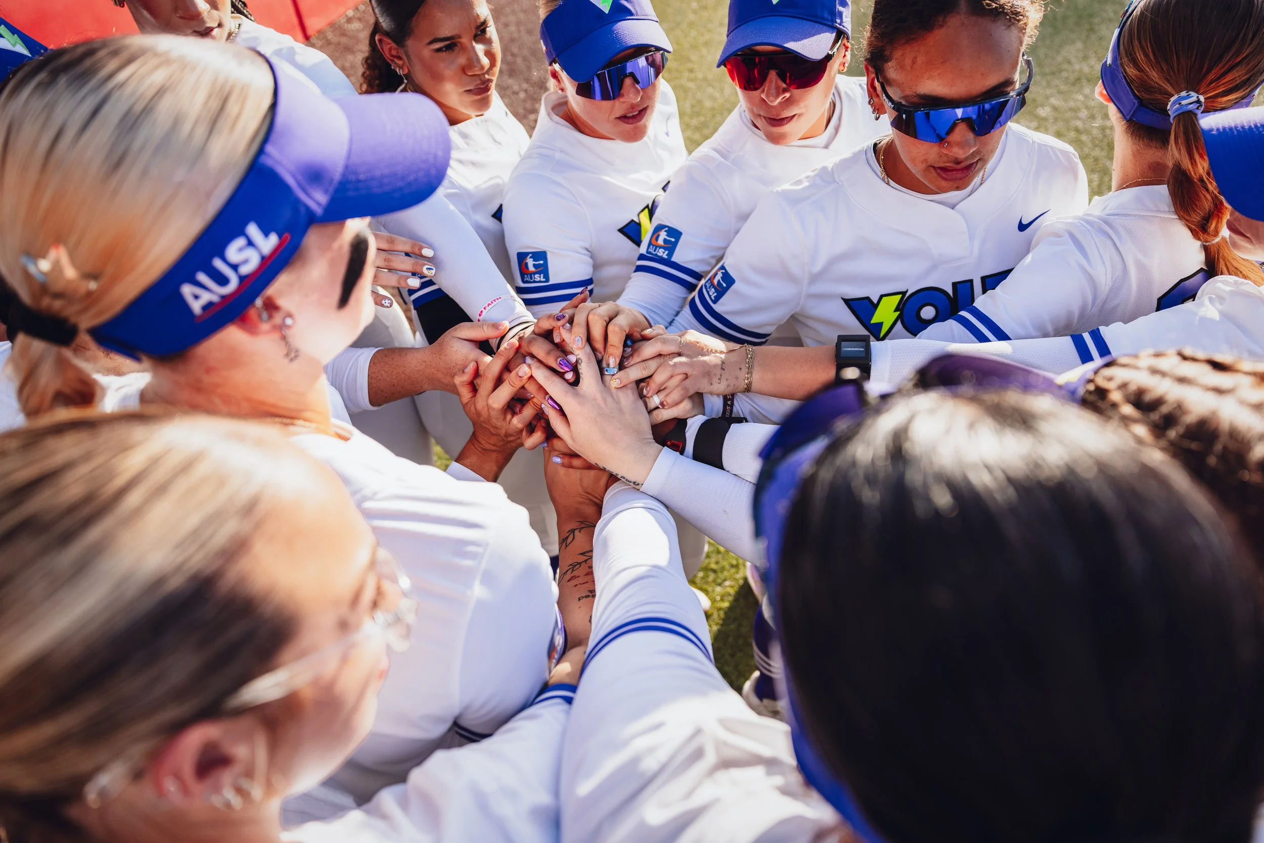 Women athletes in white jerseys and blue hats huddle together with their hands stacked in the center on a grassy field, preparing for a team cheer.