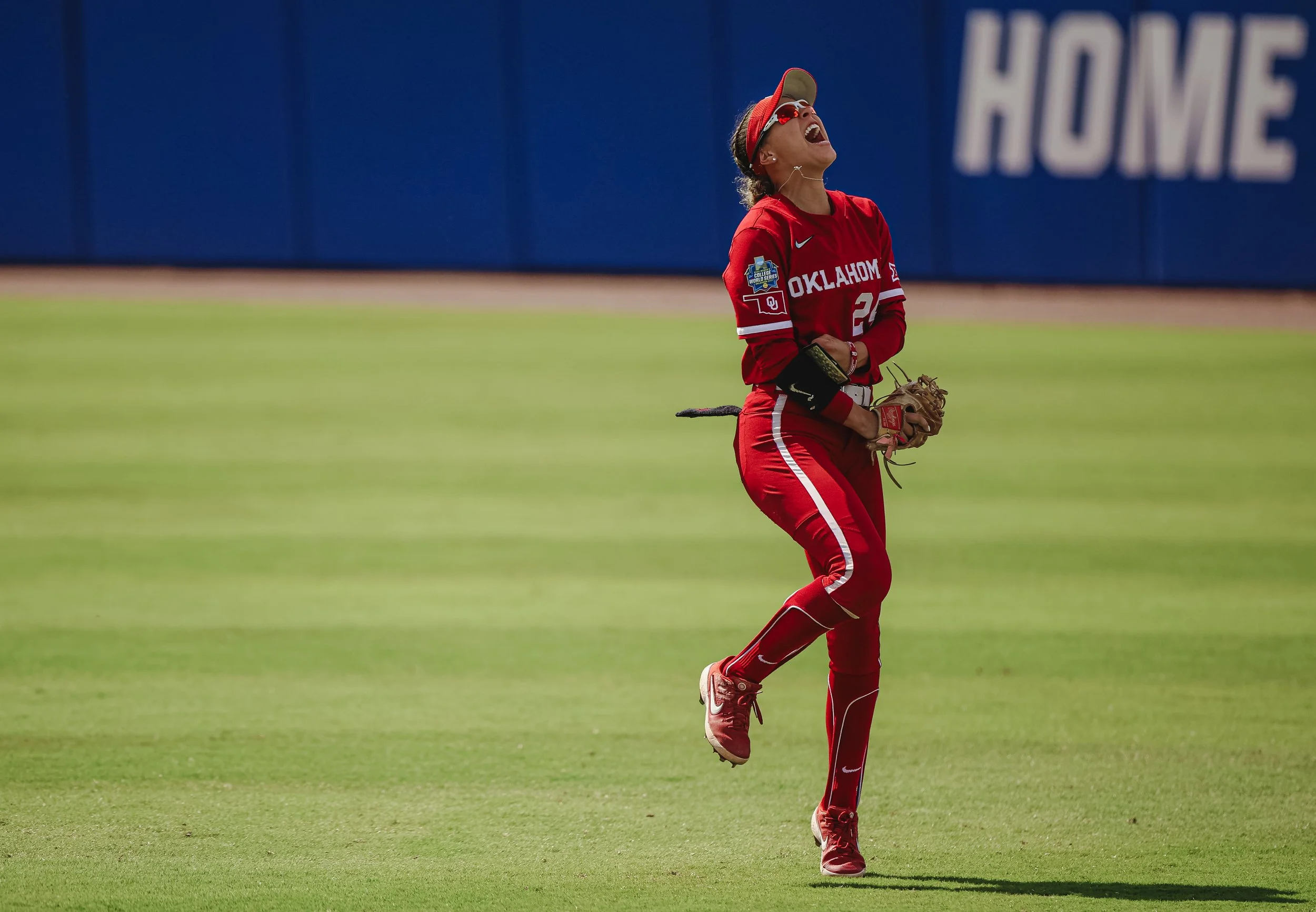 A female softball player in red Oklahoma team uniform with cap, sunglasses, and glove, laughing on the field.