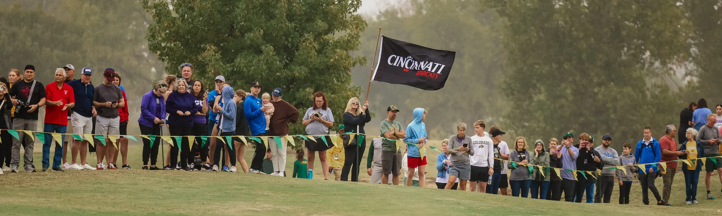 Crowd of people standing behind a yellow and green striped barrier at a scenic outdoor event, with some holding phones and a woman waving a black flag with 'CINCINNATI BEARCATS' written on it, amid a background of trees and a slightly foggy sky.