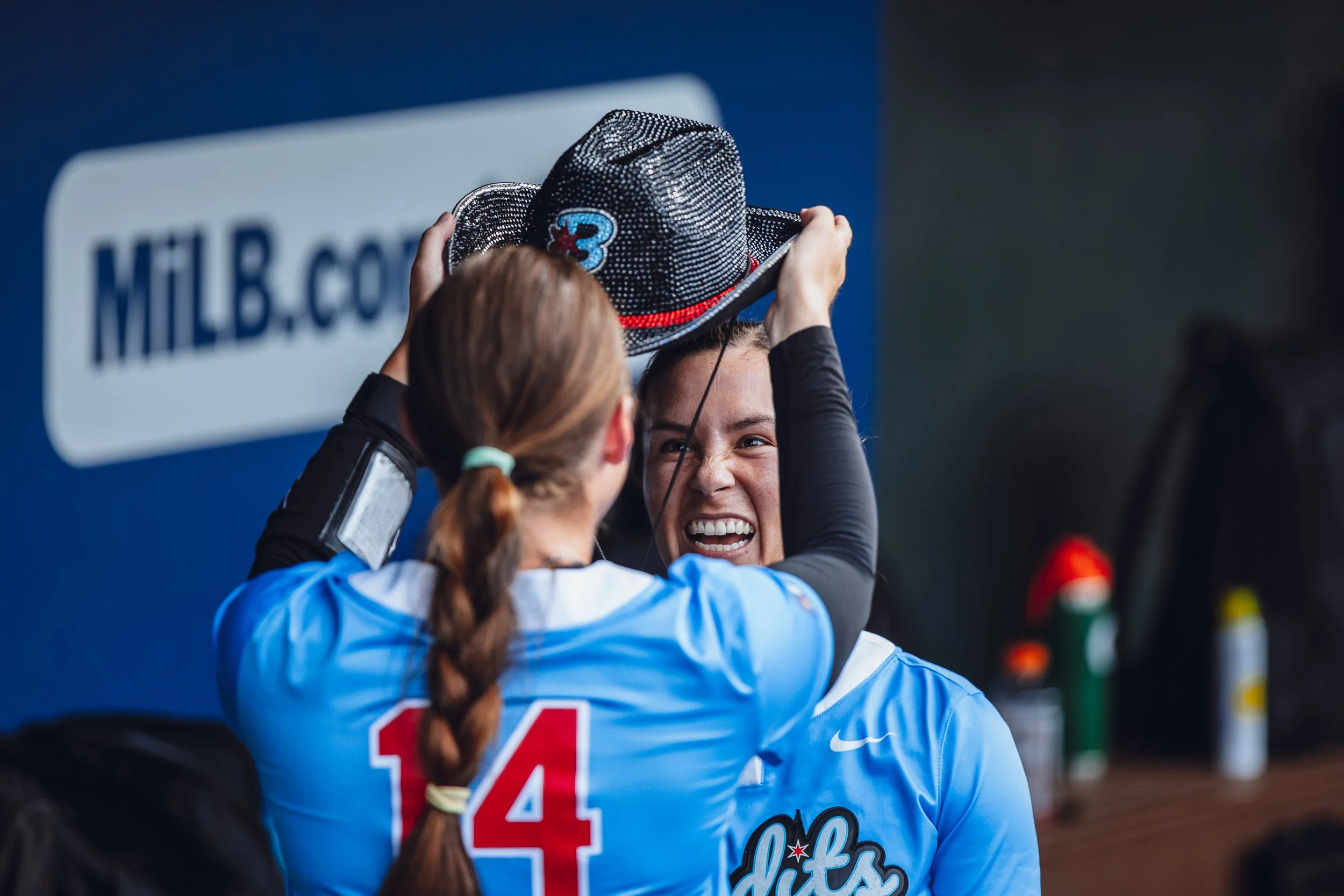 Two female softball players, one wearing a blue jersey with the number 14, are sharing a joyful moment as one helps the other put on a black sparkly cowboy hat. They are in the dugout with a sign that reads 'MLB.com' in the background.