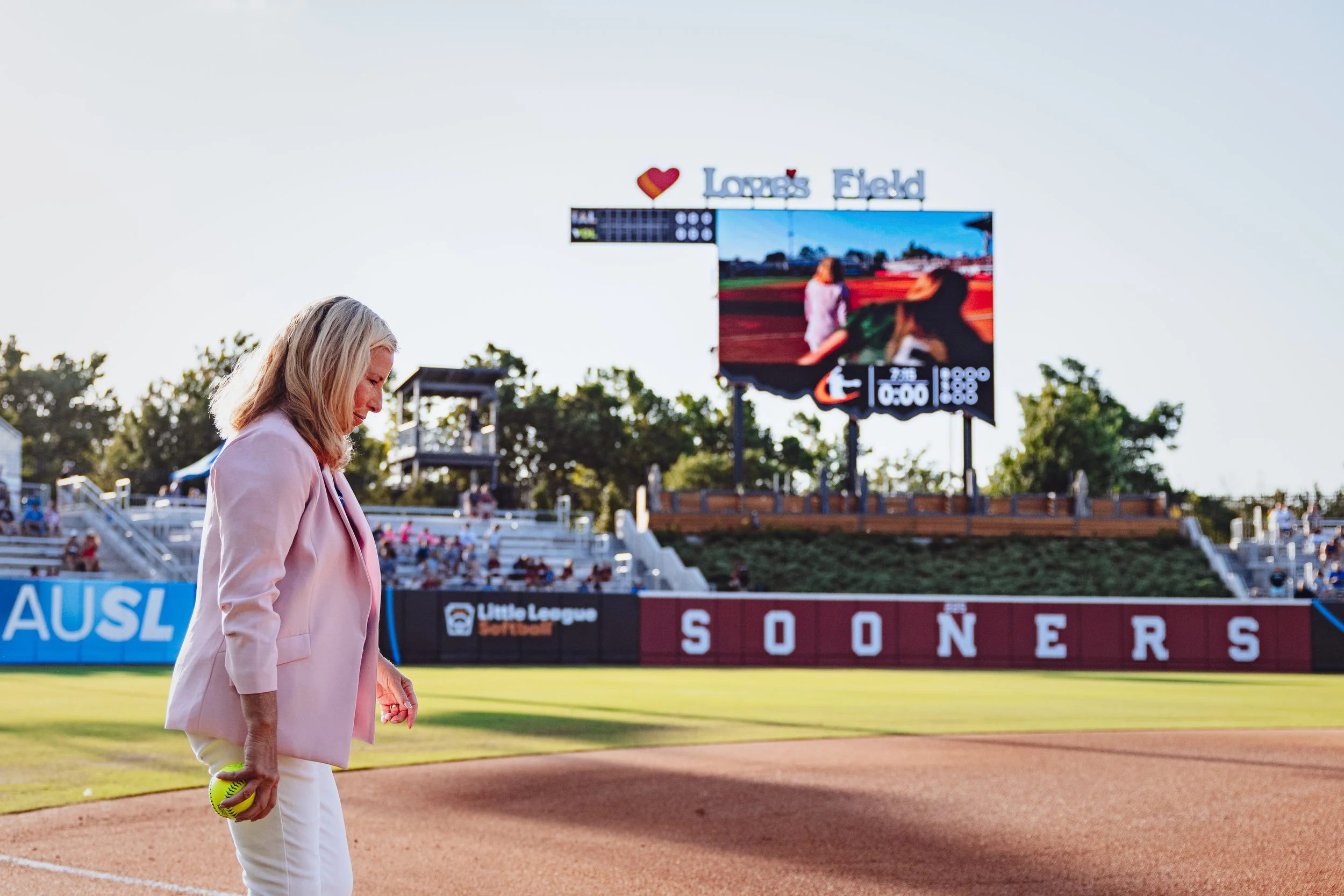 A woman in a pink blazer holding a softball walking on a baseball or softball field with a scoreboard and empty bleachers in the background.