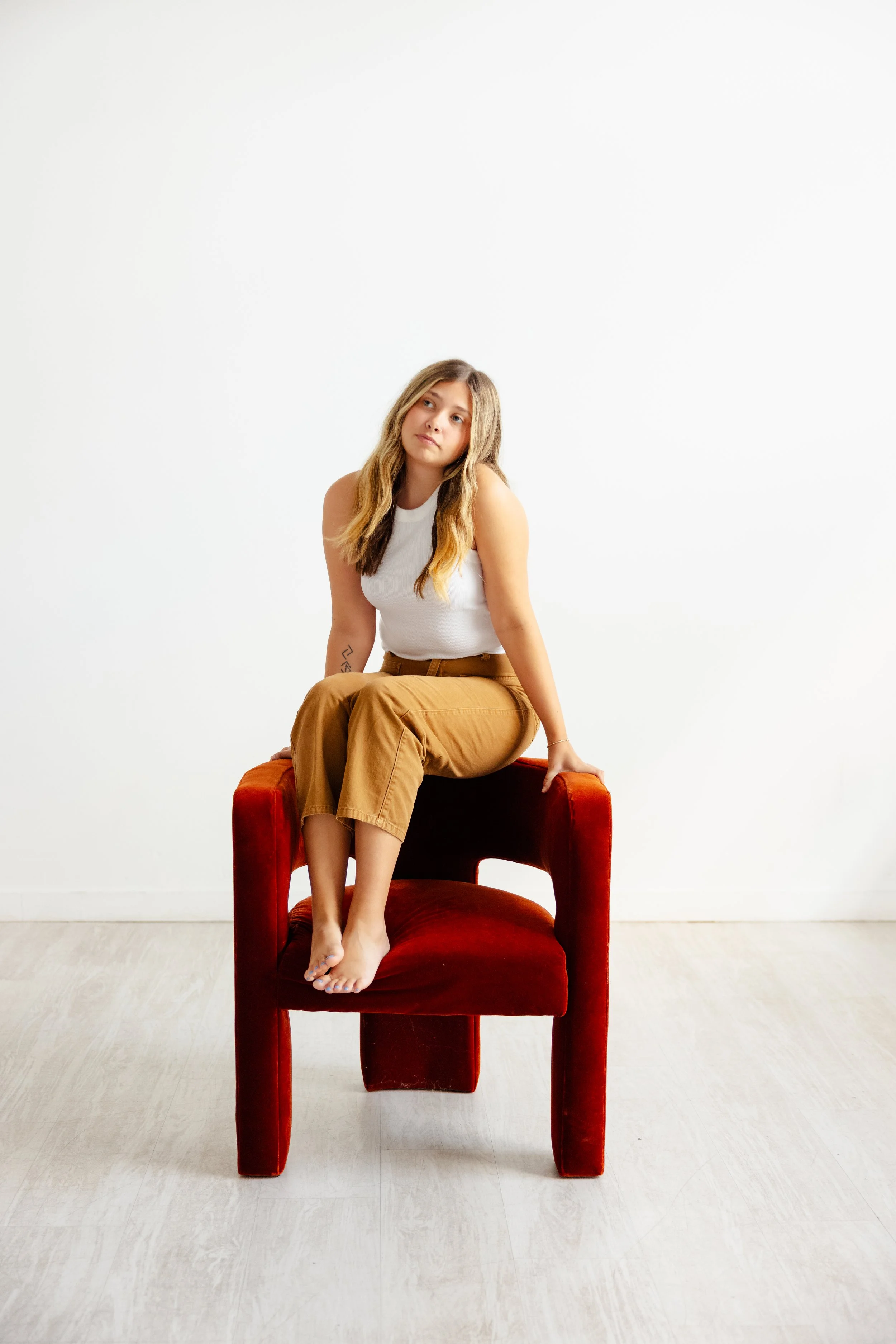 A young woman with long wavy hair wearing a white sleeveless top and brown pants, sitting barefoot on a red velvet chair with her legs crossed and looking to her left, against a plain white wall background.