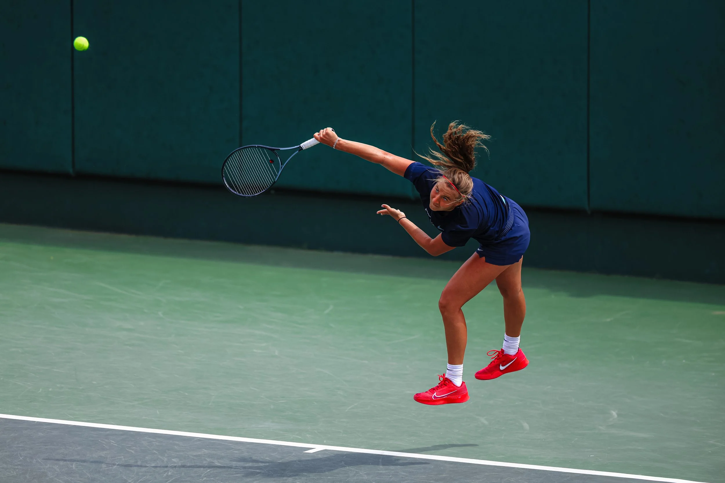 Female tennis player wearing a navy-blue outfit and red tennis shoes hitting a backhand shot on a green tennis court.