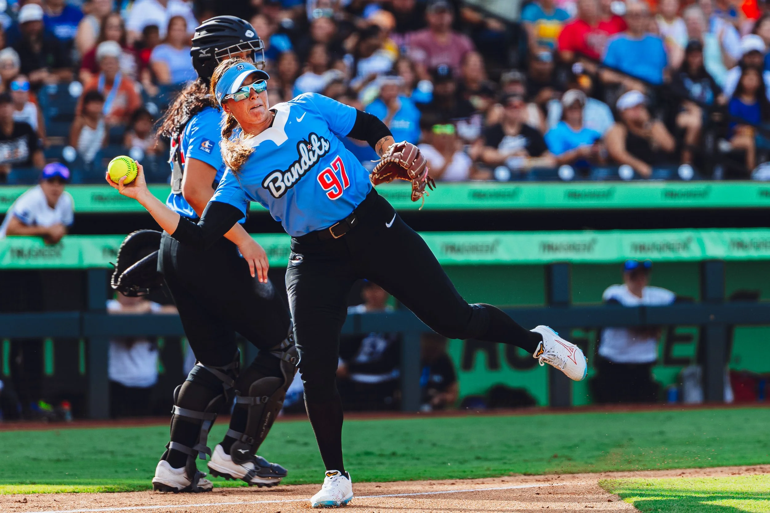 A woman in a blue jersey and sunglasses is pitching a softball during a game. An umpire behind her is in position, and there is a crowd of spectators in the background.