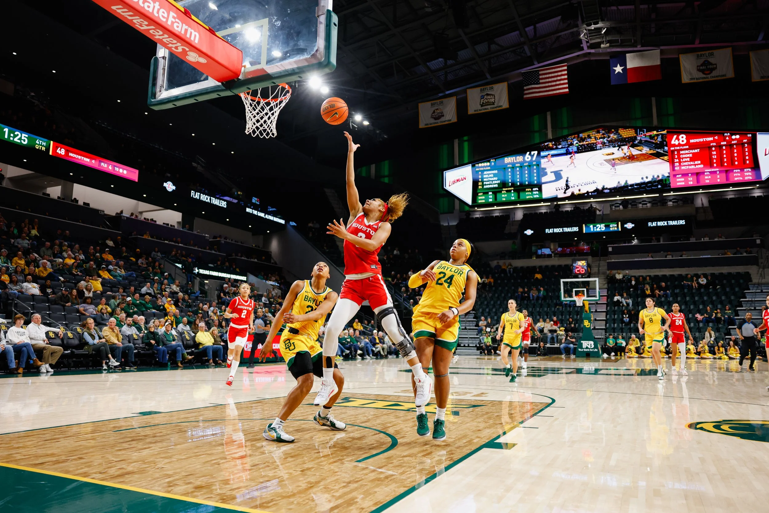 A women's basketball game between Houston and Baylor, with players on the court, one player in red jumping to score or block, and others in yellow defending; audience seated in the stands; scoreboard displaying Houston leading Baylor.