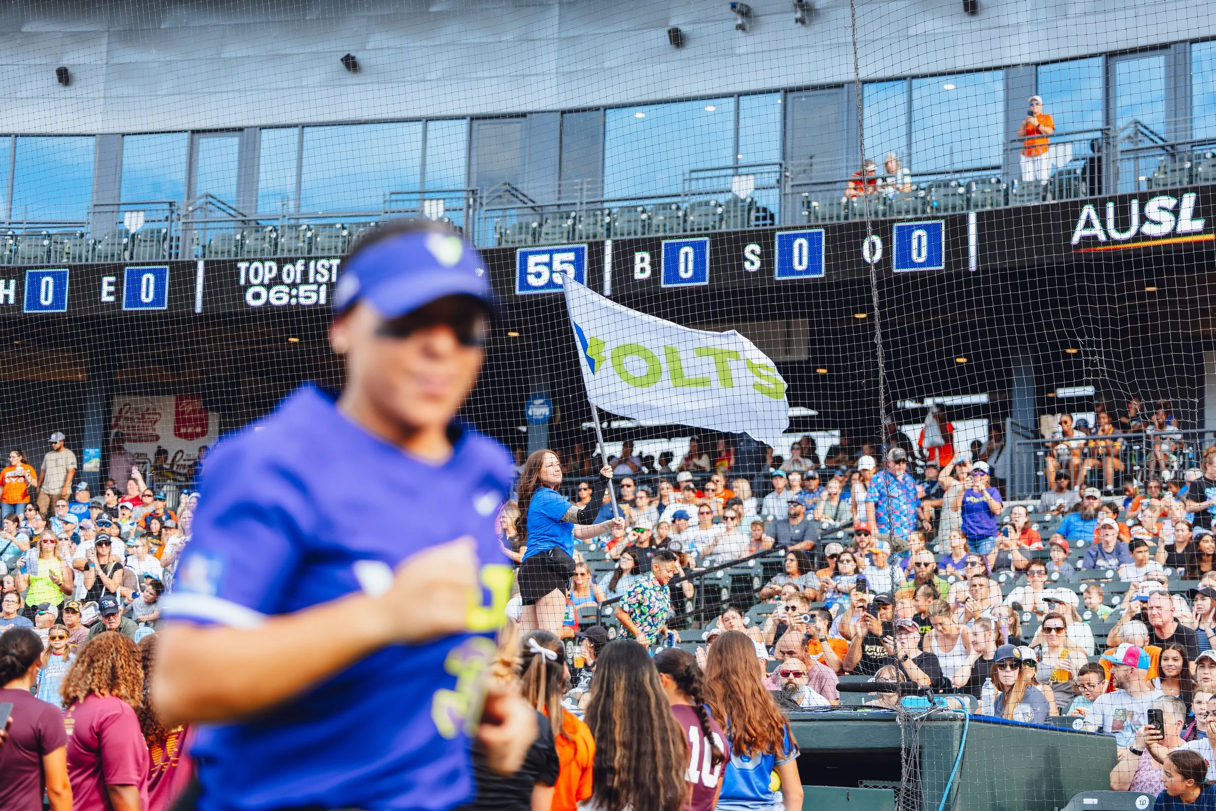 Cricket stadium crowd with fans, a woman waving a 'VOLT'S' flag, and a blurred cricket player in the foreground.