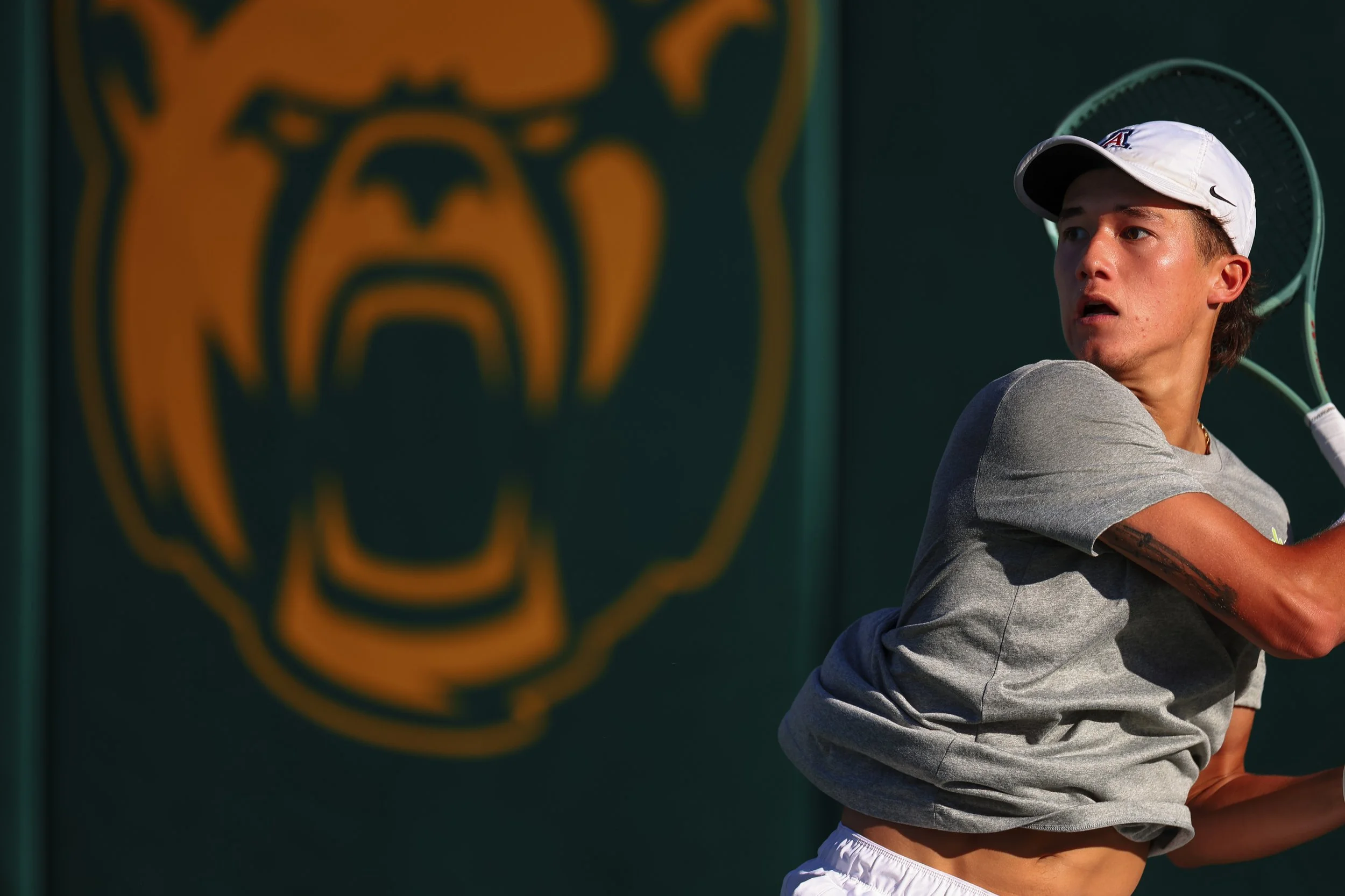 A tennis player in a gray shirt and white cap preparing to hit a shot on the tennis court, with a blurred tiger logo in the background.