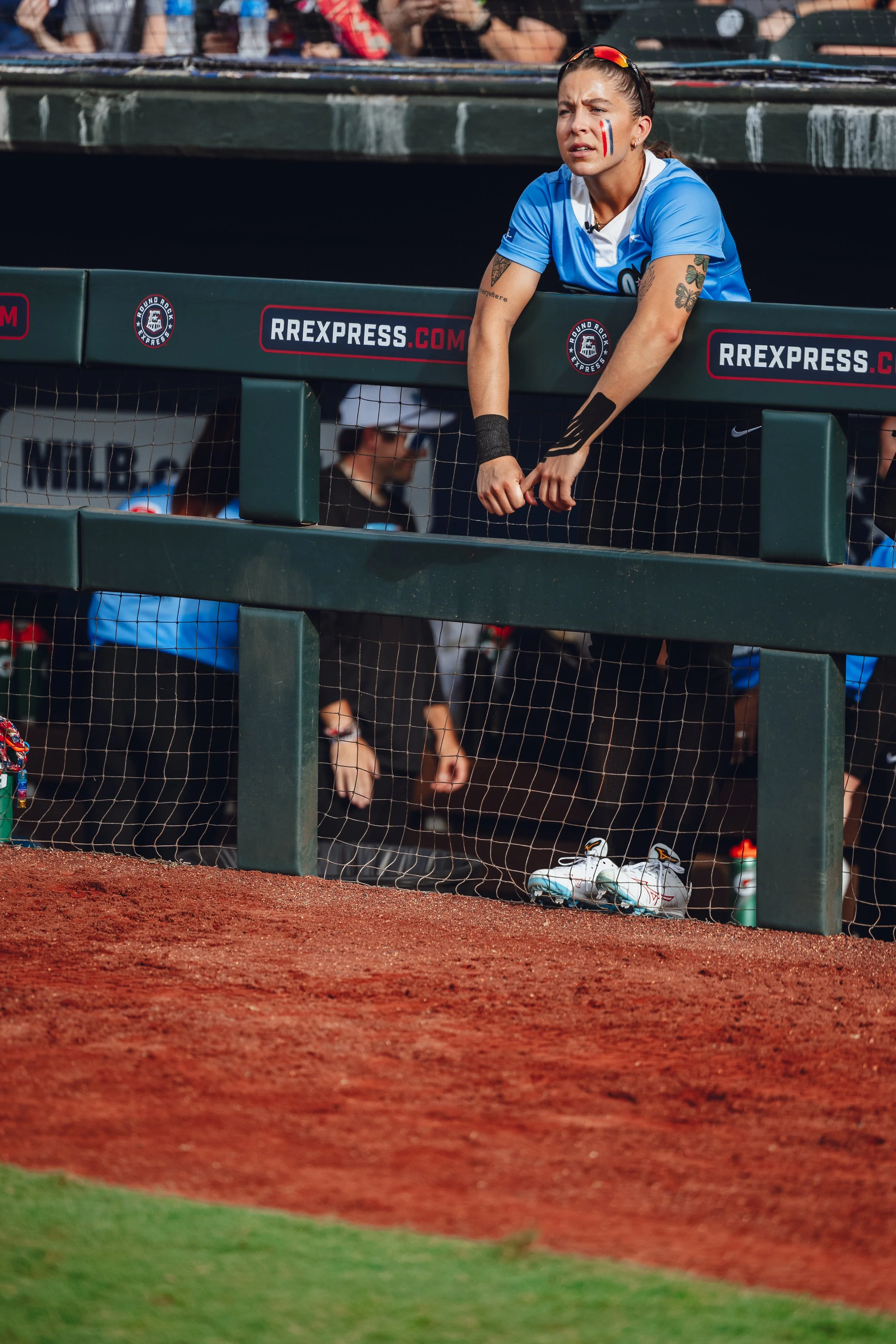 A female soccer player in a blue uniform with face paint leaning on a railing during a game, with her white cleats on the ground and a water bottle nearby.