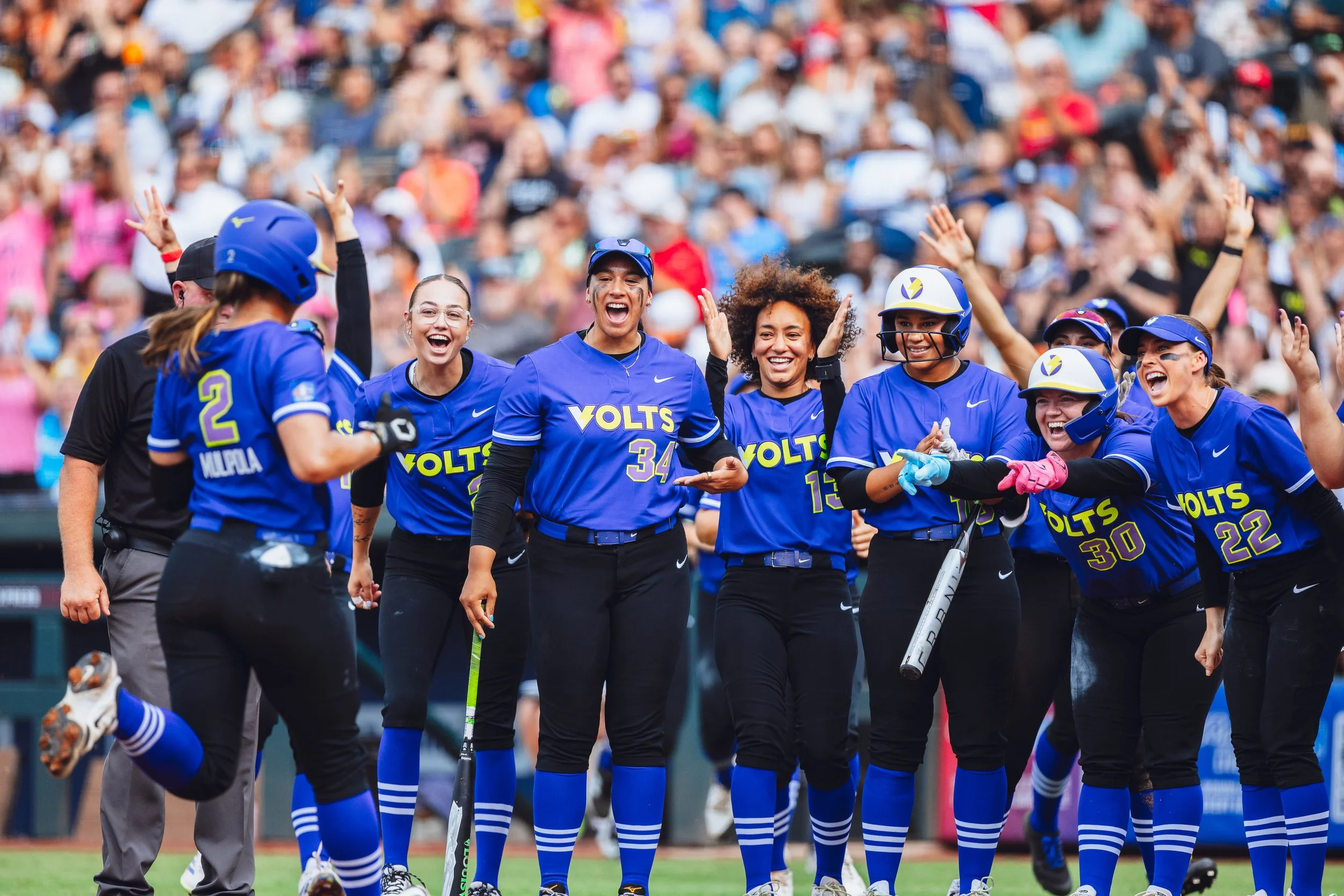 Softball team in blue uniforms celebrating on the field with smiling and cheering players, some with helmets, in front of a crowd of spectators.