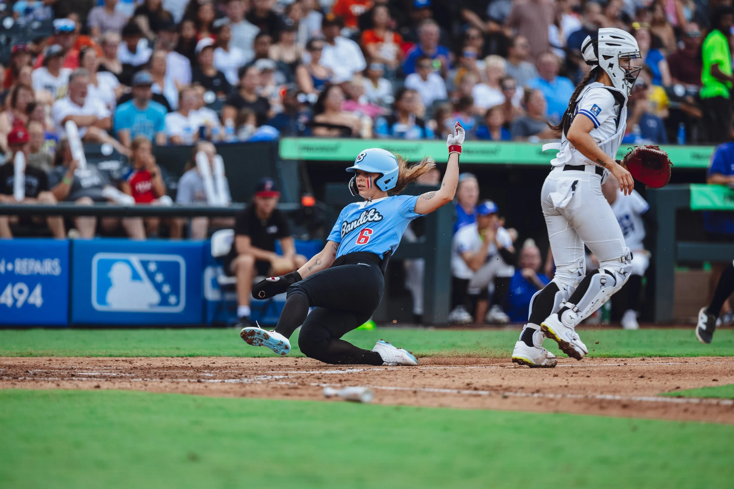 A woman sliding into home plate during a baseball game, wearing a blue jersey with the number 6, and a catcher in white gear is nearby on the field with a crowd watching.