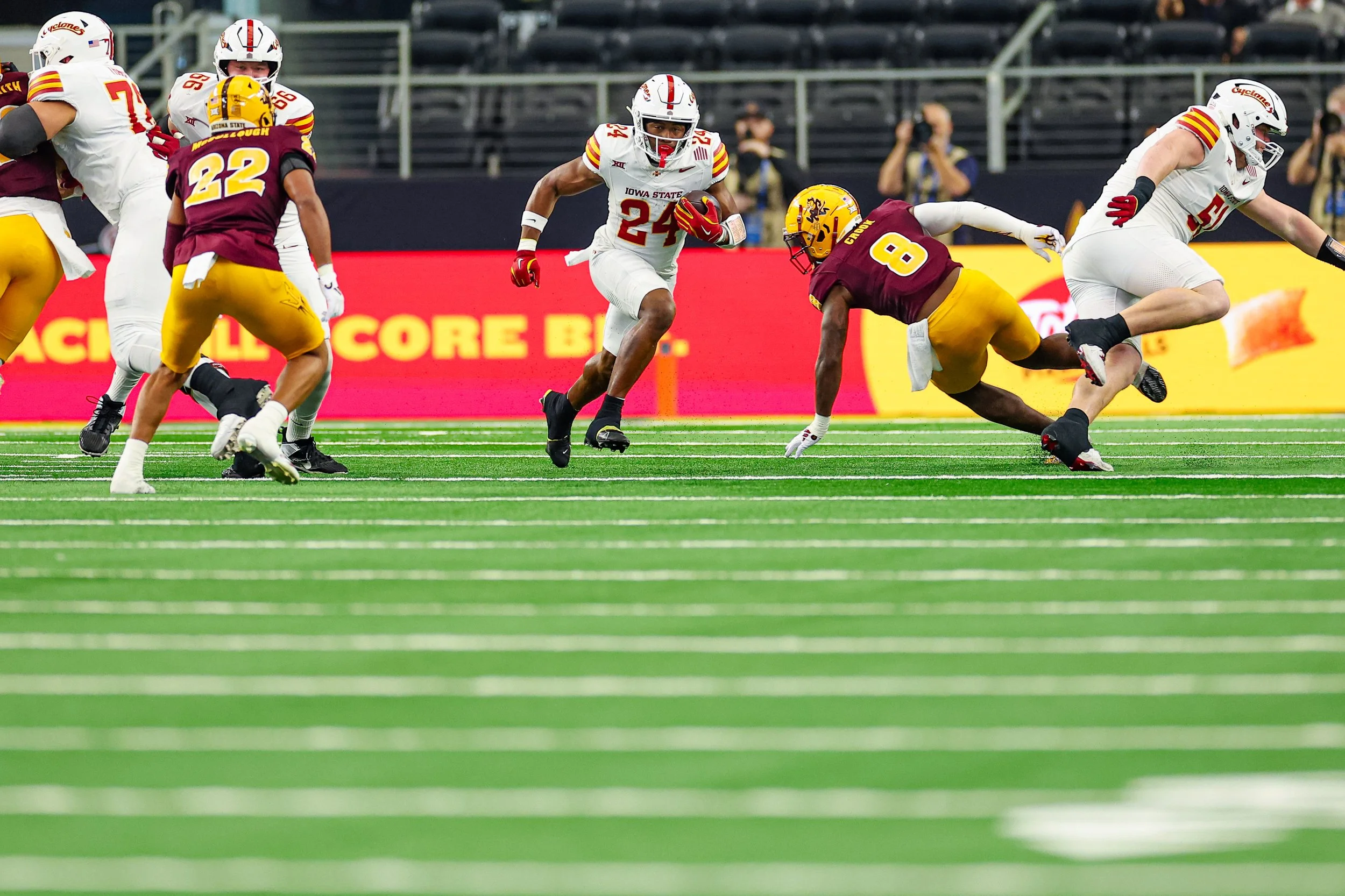 An American football game with players from Iowa State in white uniforms and opponents in maroon and gold uniforms. One player in white is running with the ball, and players in maroon are tackling and blocking on the field with green turf and white y