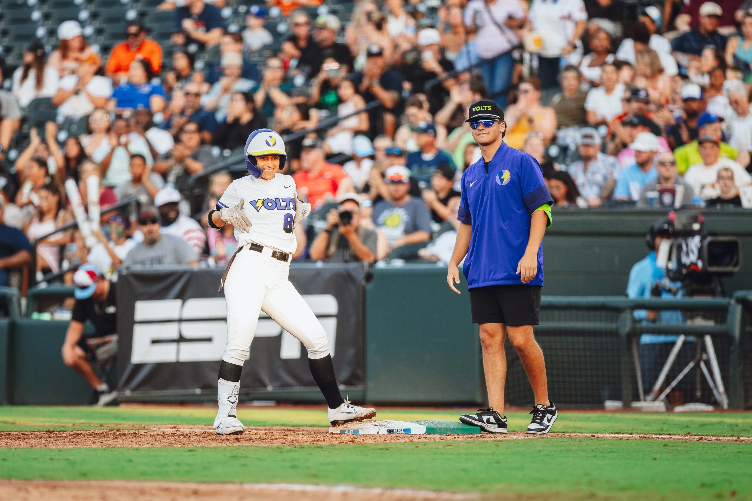 A woman in a baseball uniform standing on a baseball base, smiling, with a man in a blue shirt and shorts standing nearby on the field. A large crowd is in the background, watching the game.