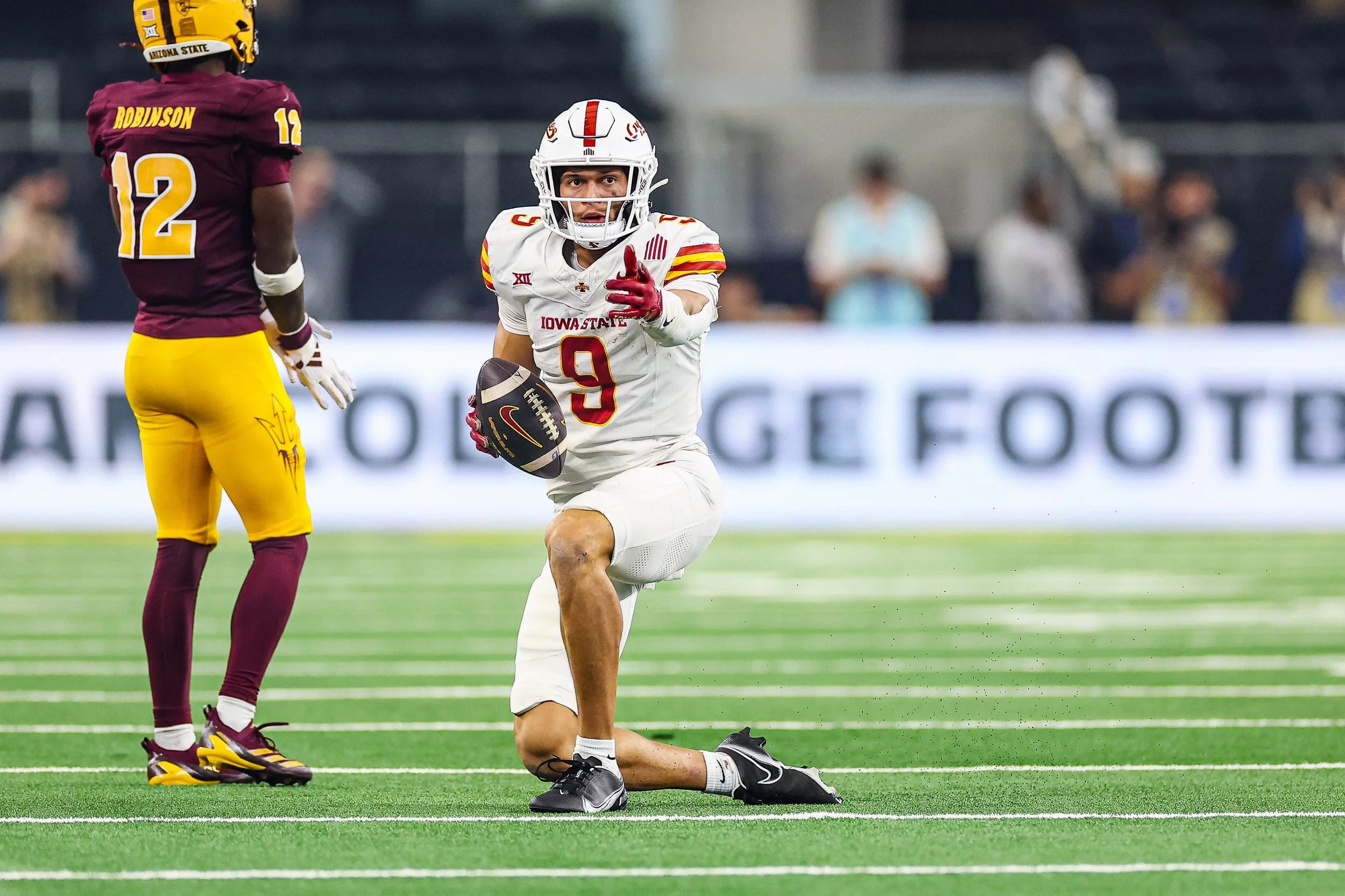A football player in a white Iowa State uniform kneeling on one knee on a football field. The player is holding a football and pointing forward. Another player in a maroon and yellow uniform is standing nearby, looking in the same direction. In the b