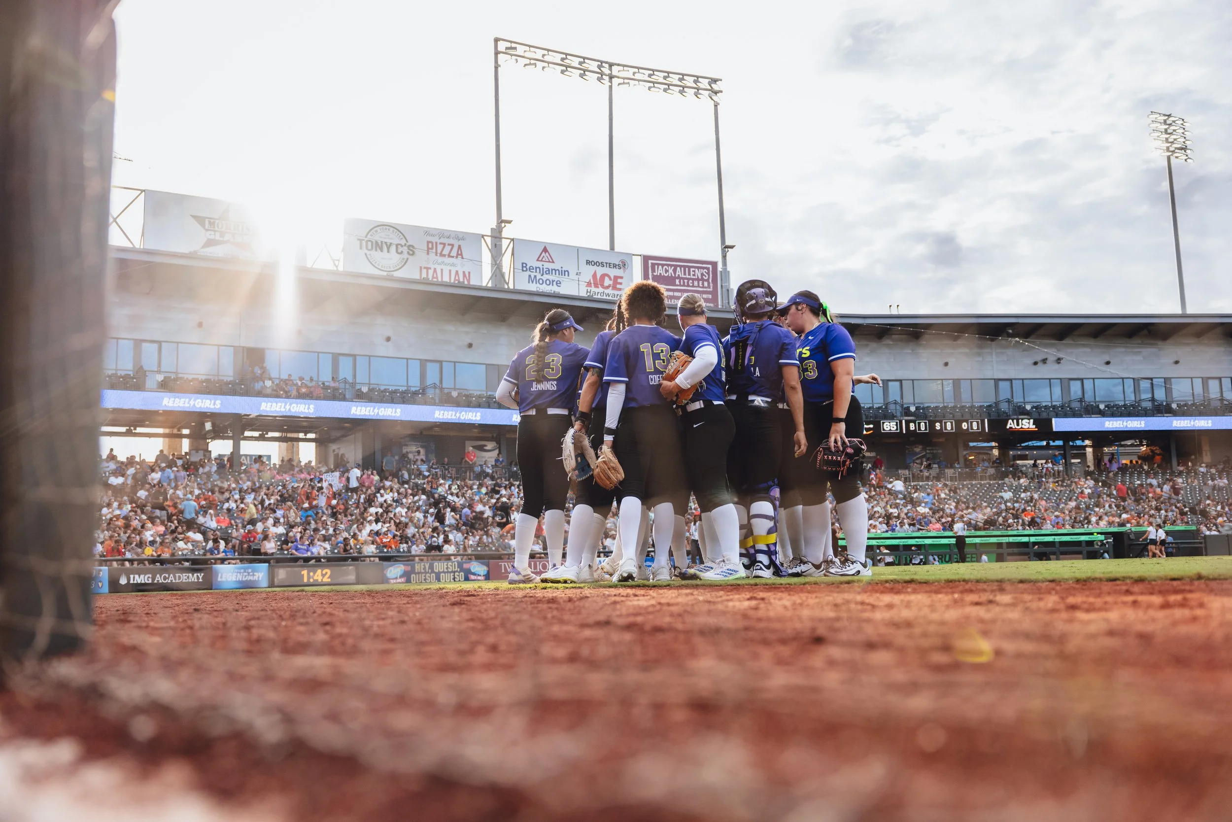 Softball team huddled together on the field during a game at a stadium with a large crowd and bright stadium lights.