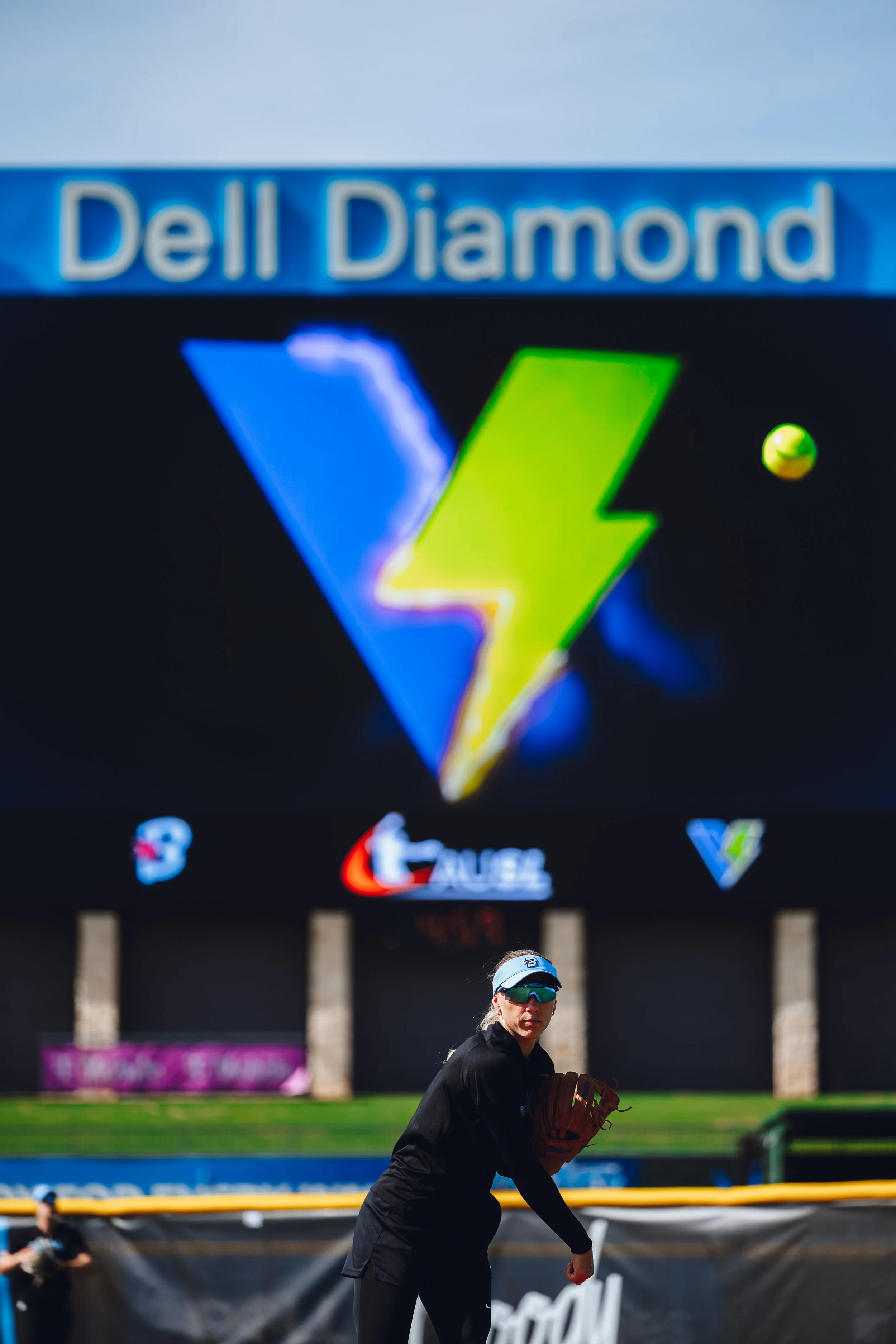 A woman in athletic clothing is preparing to throw a baseball in front of a large digital billboard at a baseball stadium.