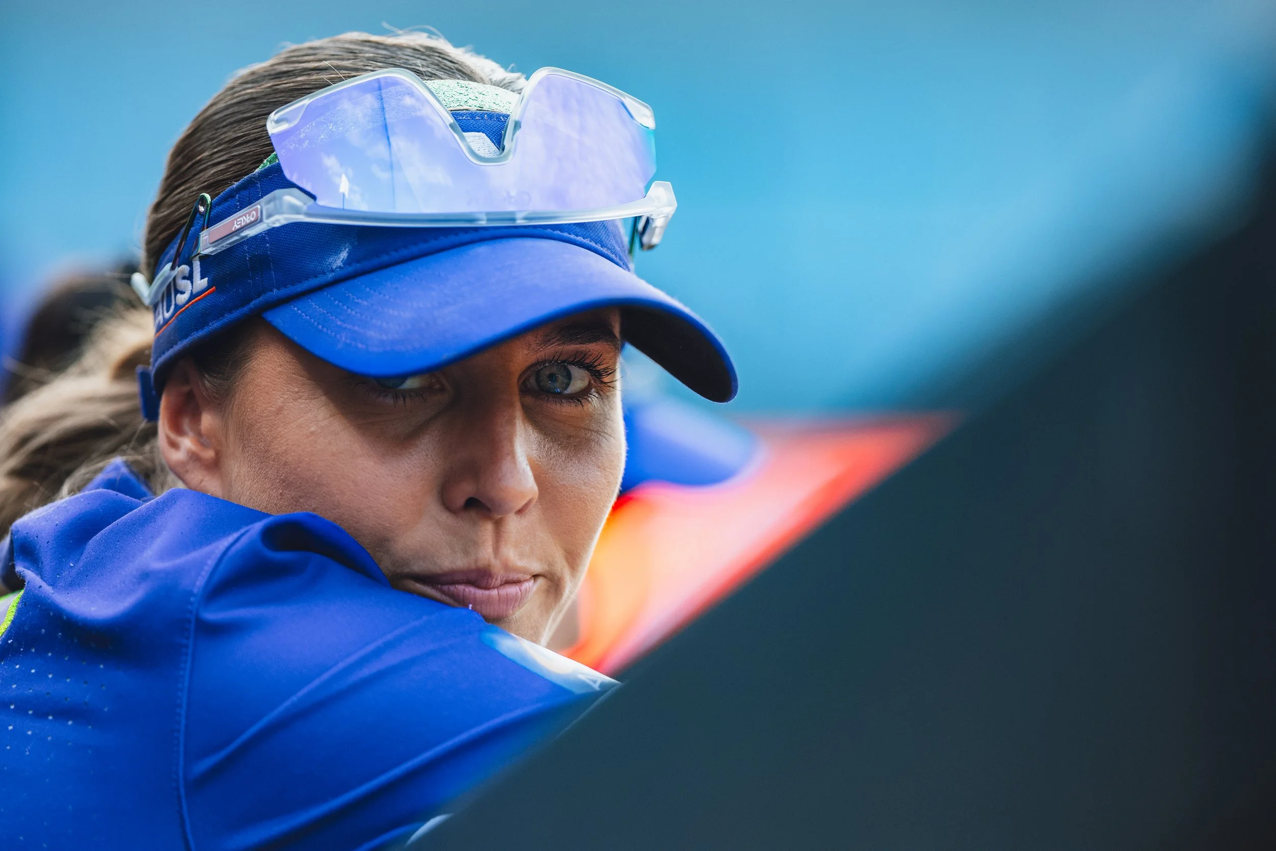 Close-up of a female athlete, wearing a blue cap with sunglasses on top, a blue jacket, and looking over her shoulder with a focused expression.