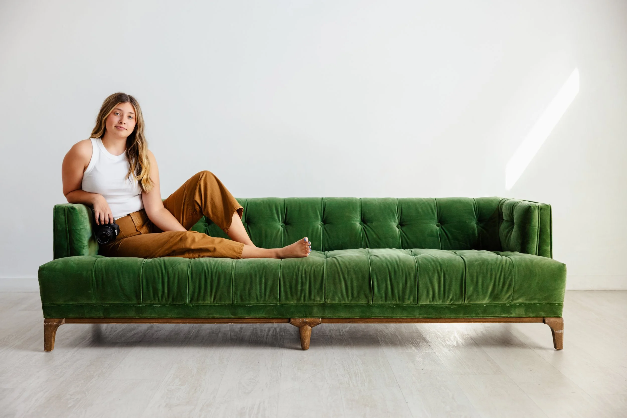 Young woman with long hair sitting on a green velvet sofa, holding a camera, in a minimalistic room with white walls and light-colored floor.